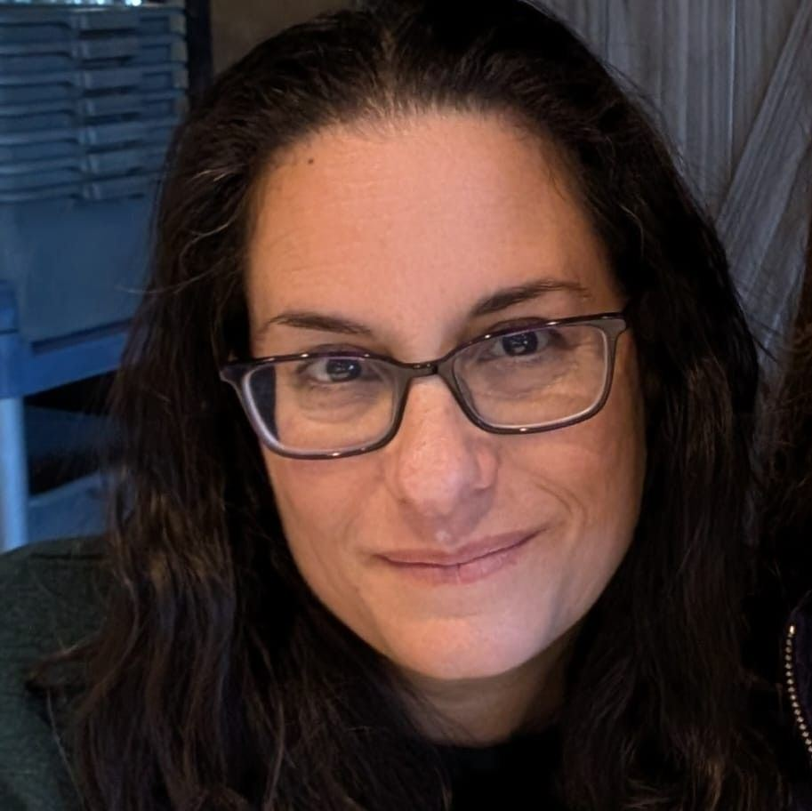 A woman with dark wavy hair, wearing glasses and earrings, smiling at the camera.