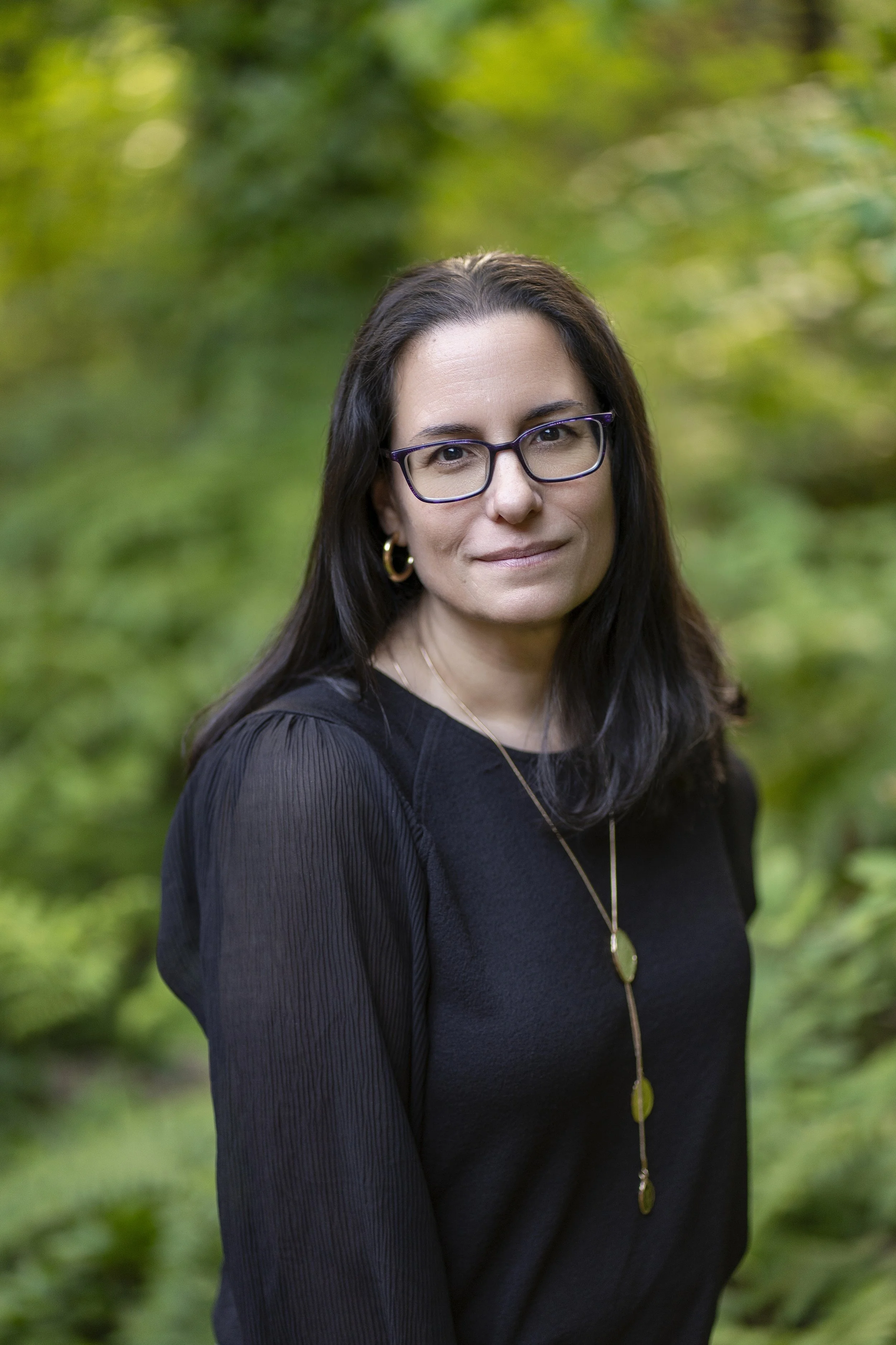 A woman with dark wavy hair, wearing glasses and earrings, smiling at the camera.