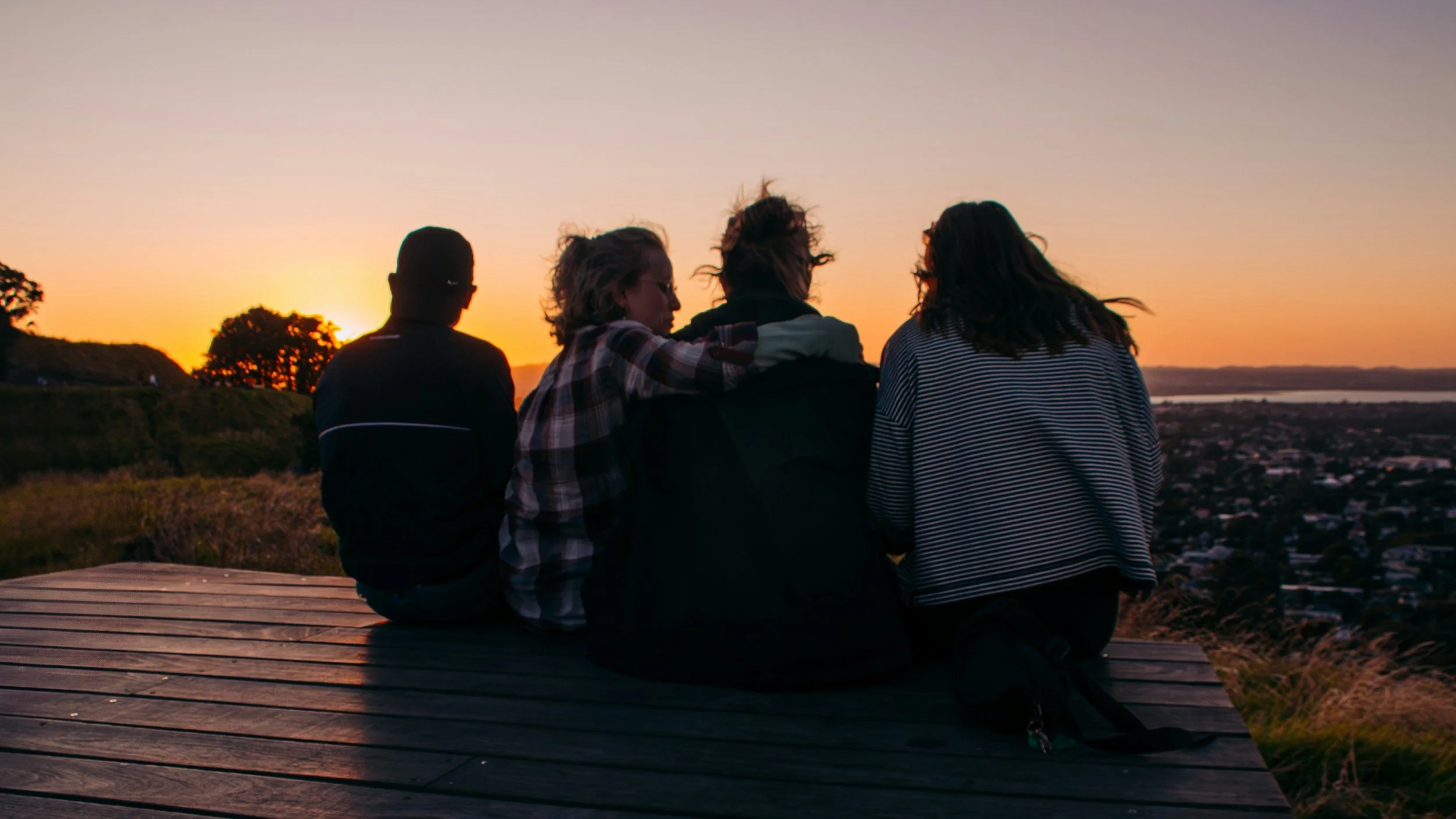 Four people sitting on a wooden platform, watching a sunset over a city and body of water, with trees and hills in the background.