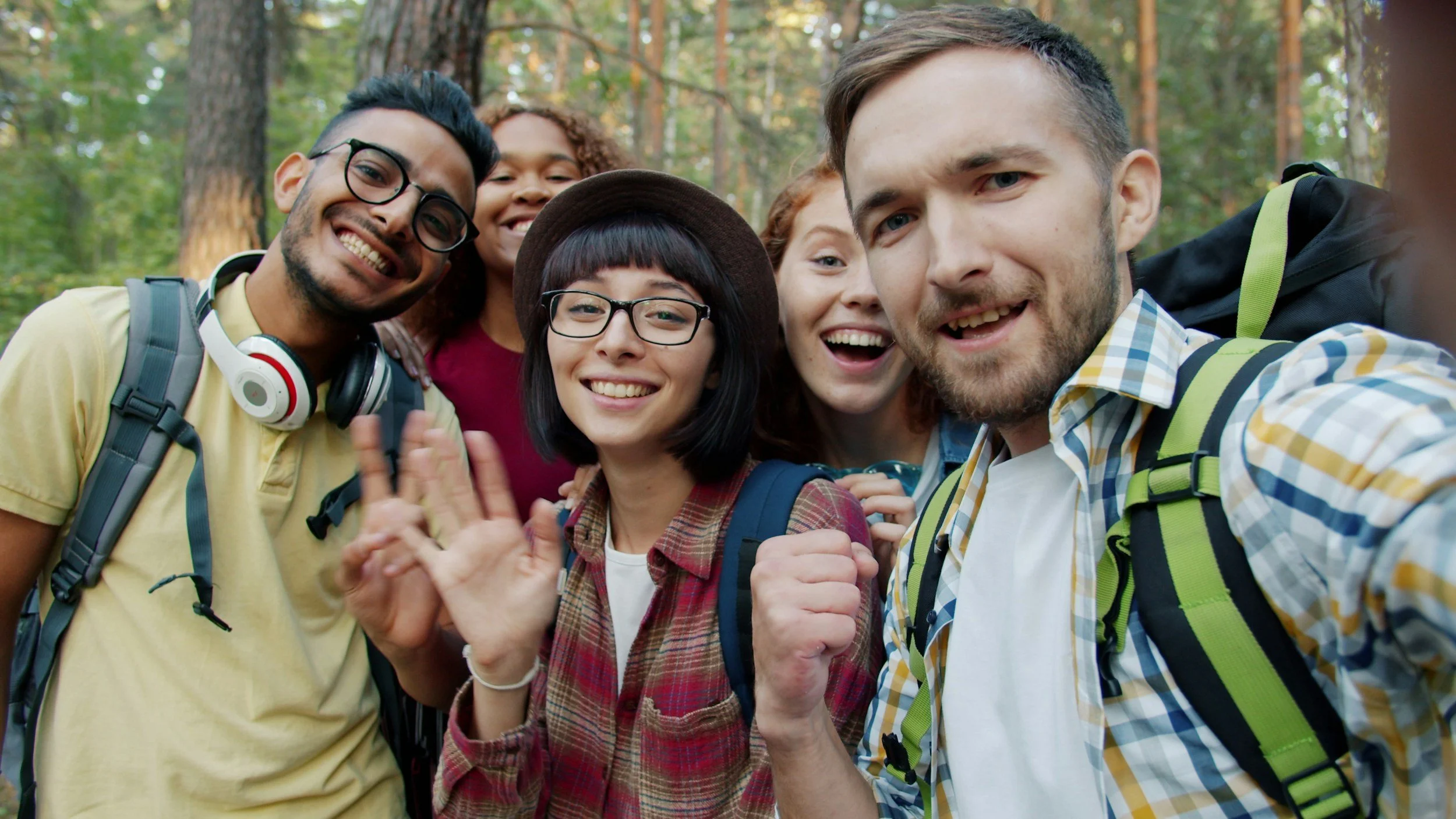 Group of six diverse young people smiling and taking a selfie in a wooded outdoor area.