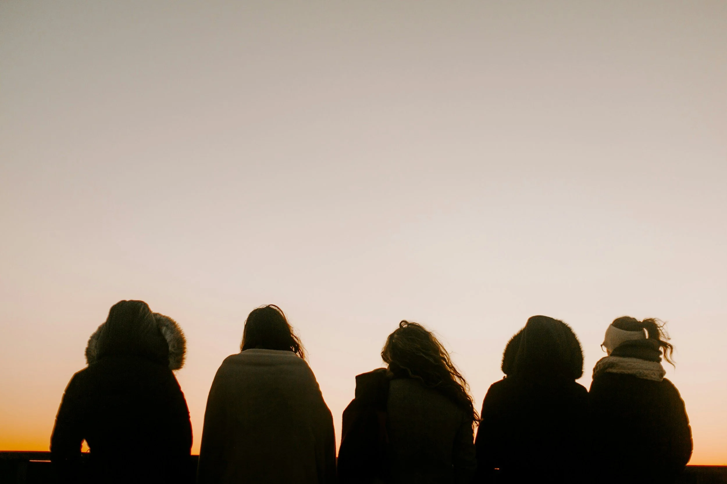 Silhouettes of five women sitting outdoors during sunset, facing away from the camera, wearing warm clothing.