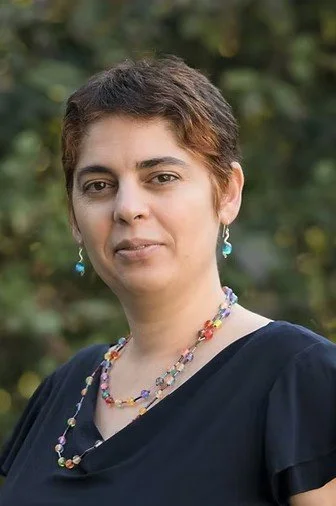 A woman with short brown hair, wearing a black top and colorful beaded jewelry, standing outdoors with blurred green foliage in the background.