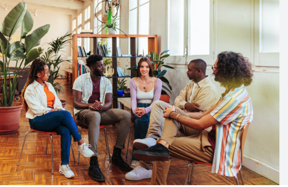 A diverse group of five people participating in a support or therapy group session in a bright, cozy room with plants and bookshelves.