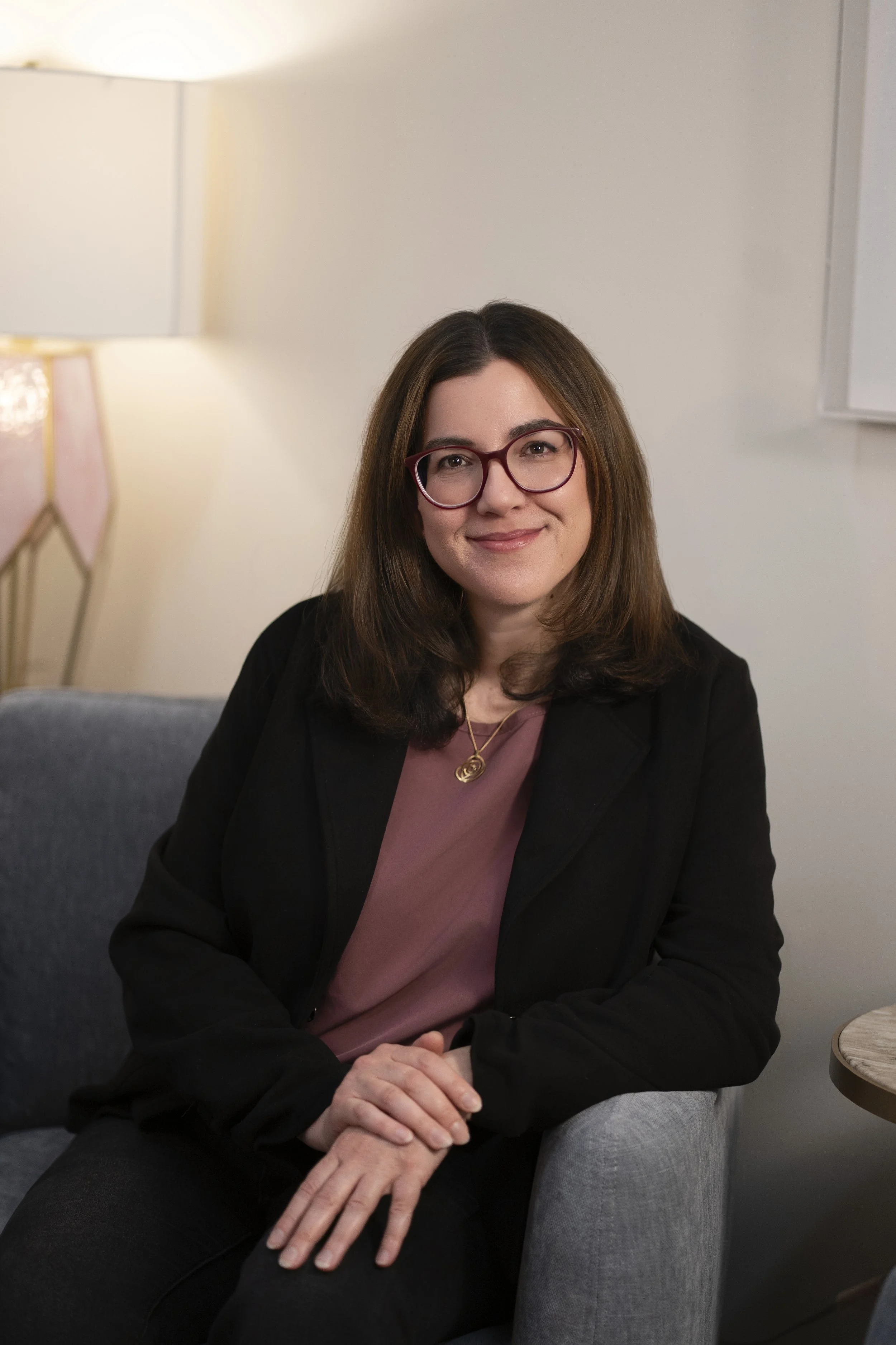 A woman with dark brown hair and glasses smiling while sitting in a white chair in an indoor setting with a window and plants in the background.