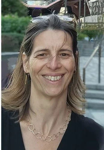 Smiling woman with shoulder-length hair, wearing earrings, a necklace, sunglasses on her head, and a black top outdoors.
