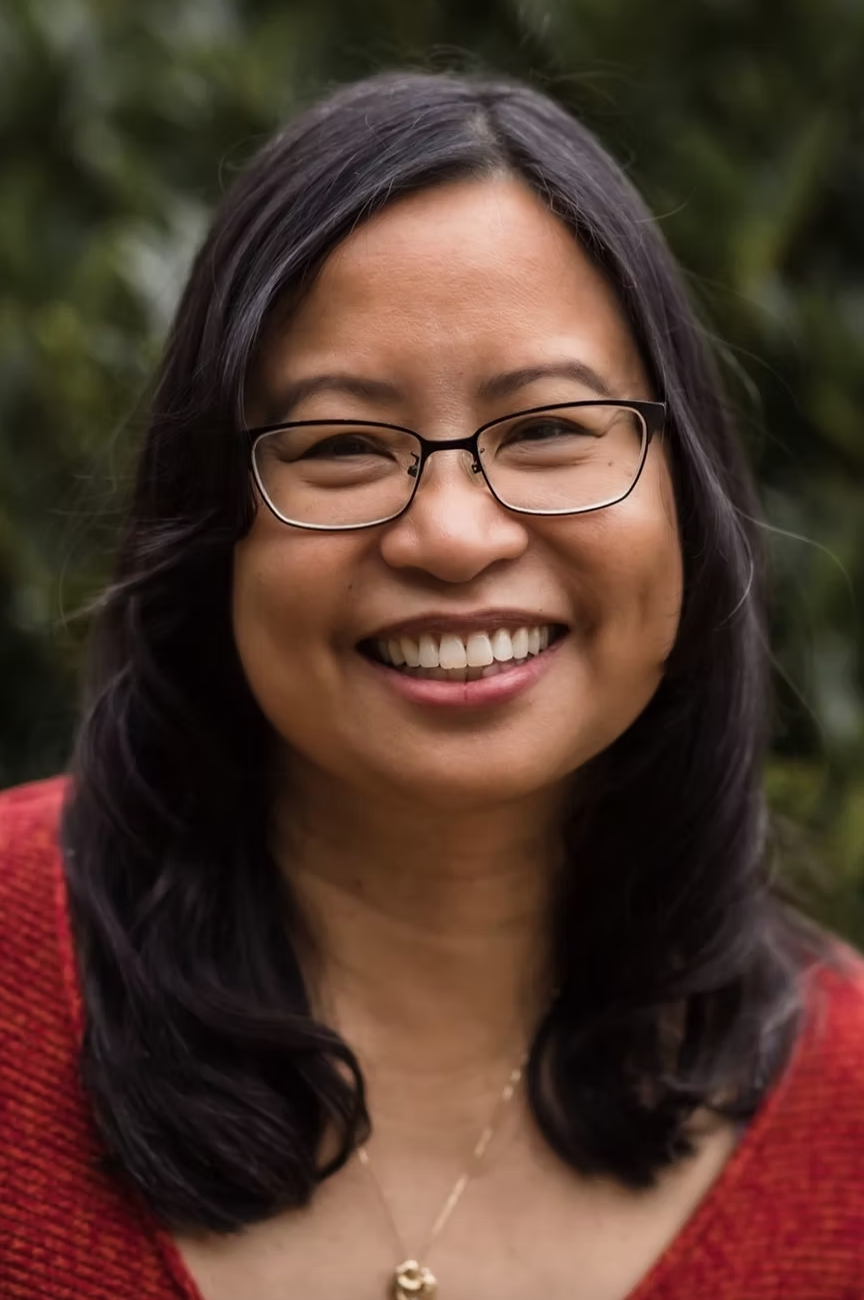 A woman with glasses smiling outdoors, wearing a red top and a gold necklace, with blurred green foliage in the background.