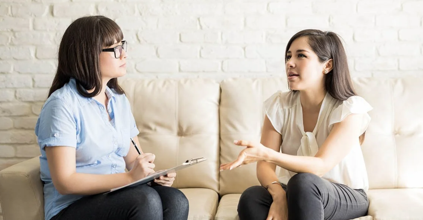 A young woman with curly hair talking to a female therapist in a clinic, sitting on a light blue sofa near a potted plant.