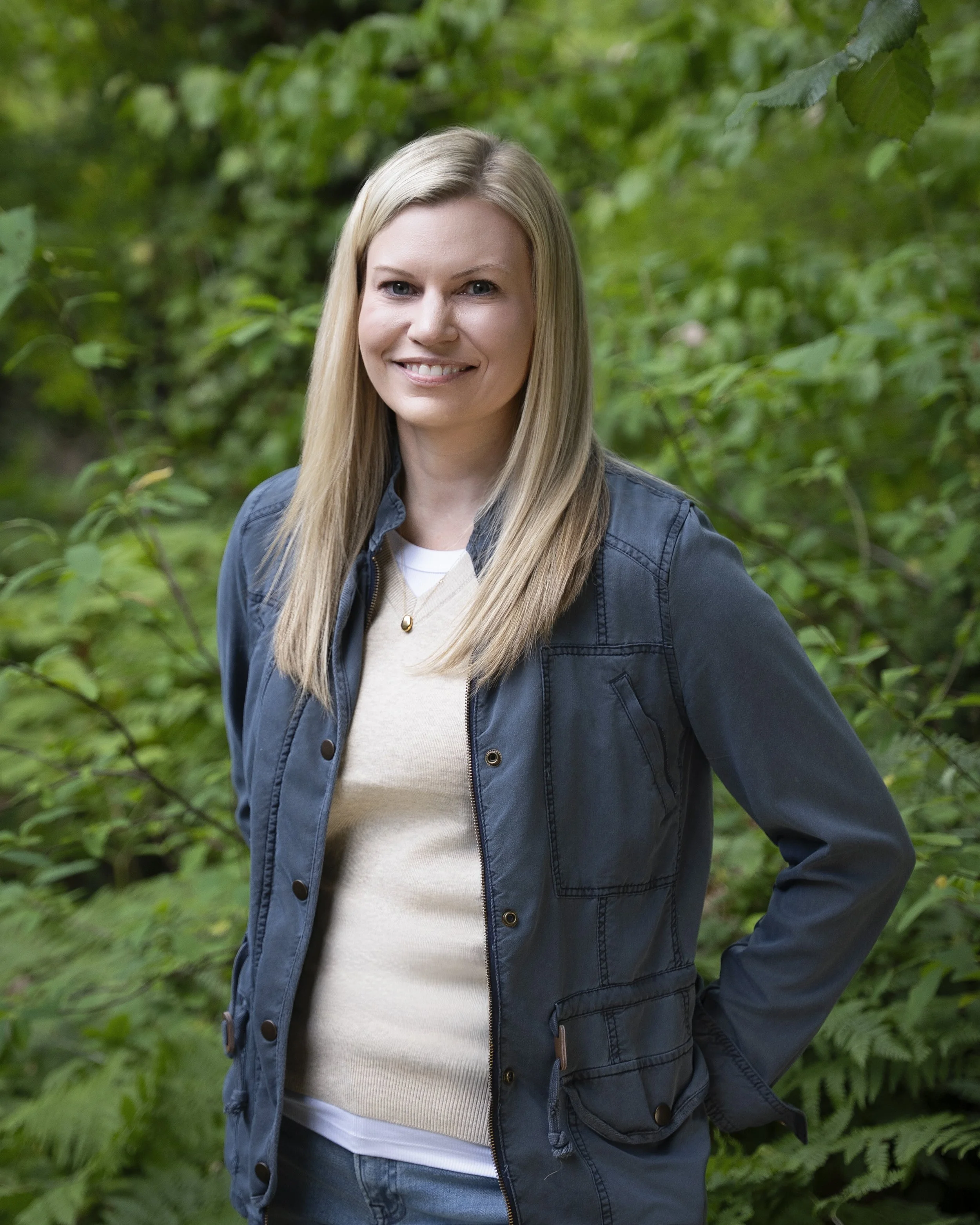 A woman with blonde hair smiling and standing against a brick wall, wearing a black knit cardigan, a teal top, a necklace with a blue pendant, and dark pants.