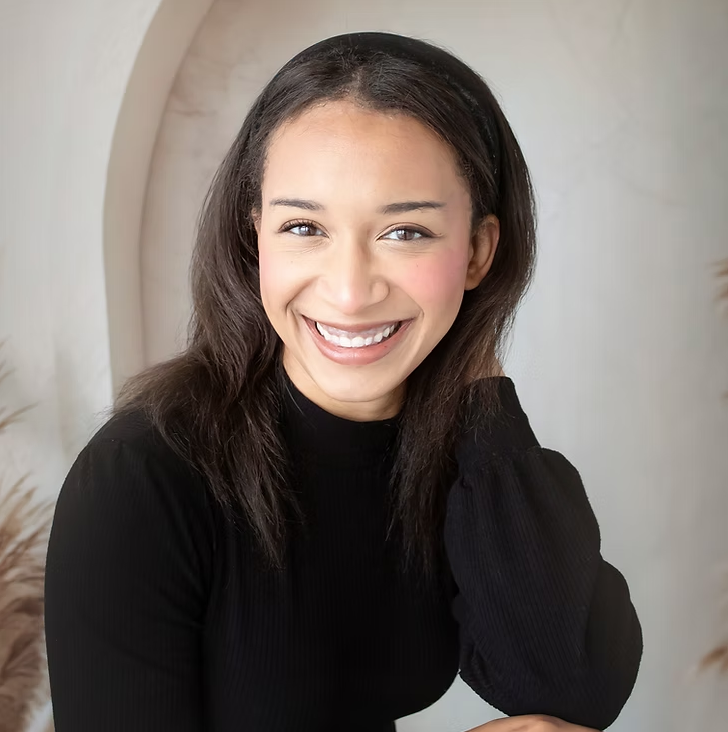 A young woman with dark hair smiling at the camera, wearing a black top, in a neutral background with some flowers.