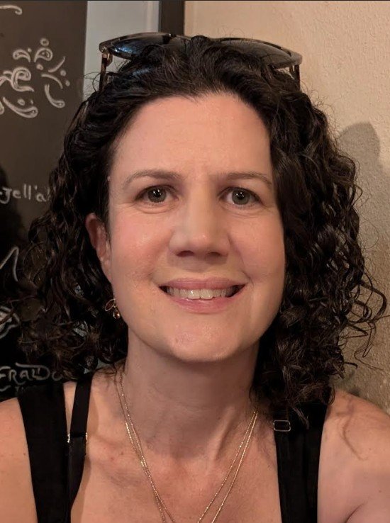 A woman with curly dark hair, wearing sunglasses on her head, smiling, with a black top and layered necklaces, standing indoors.