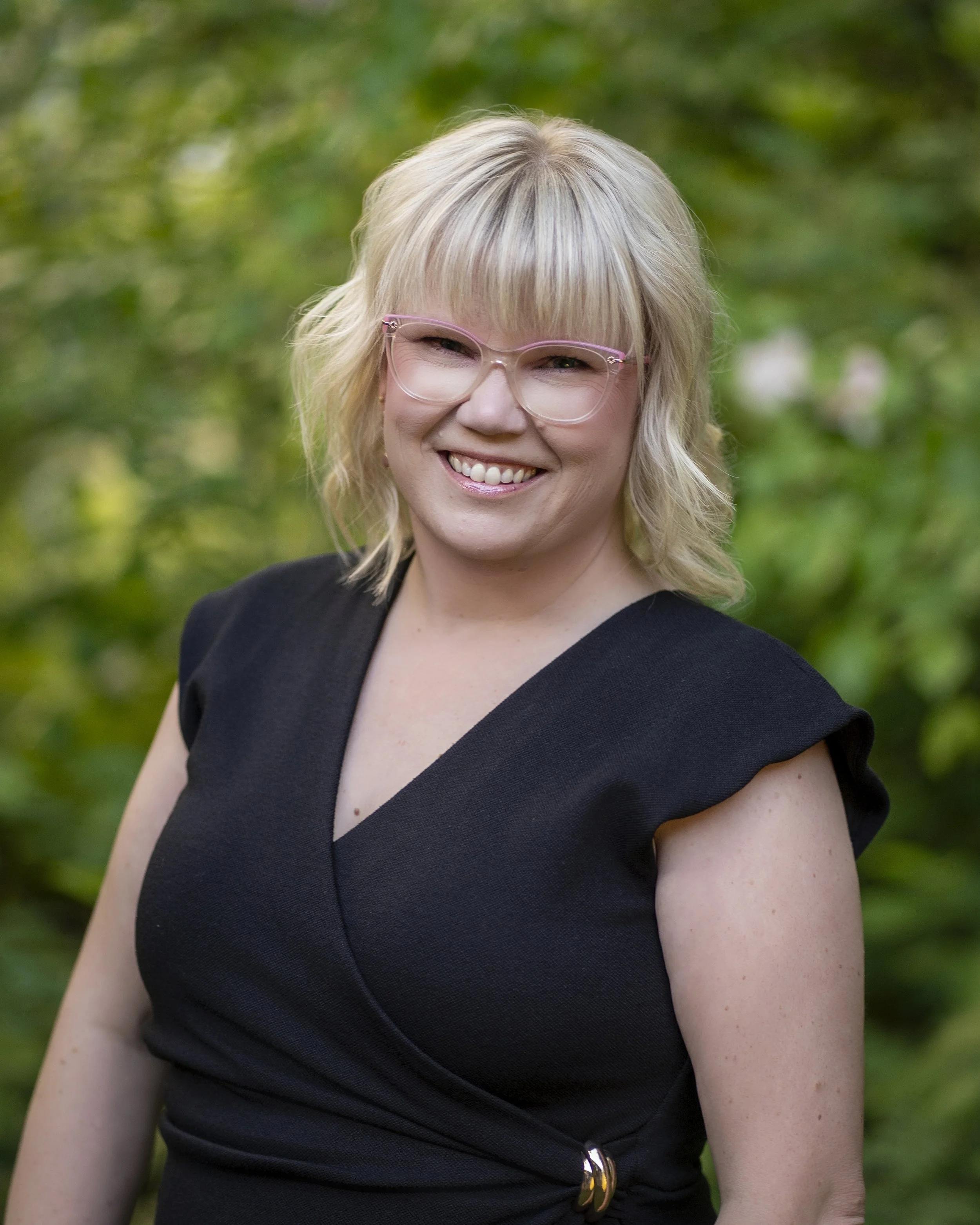 A smiling woman with blonde hair and glasses, wearing a light-colored cardigan over a black and white patterned top, standing outdoors near a body of water.
