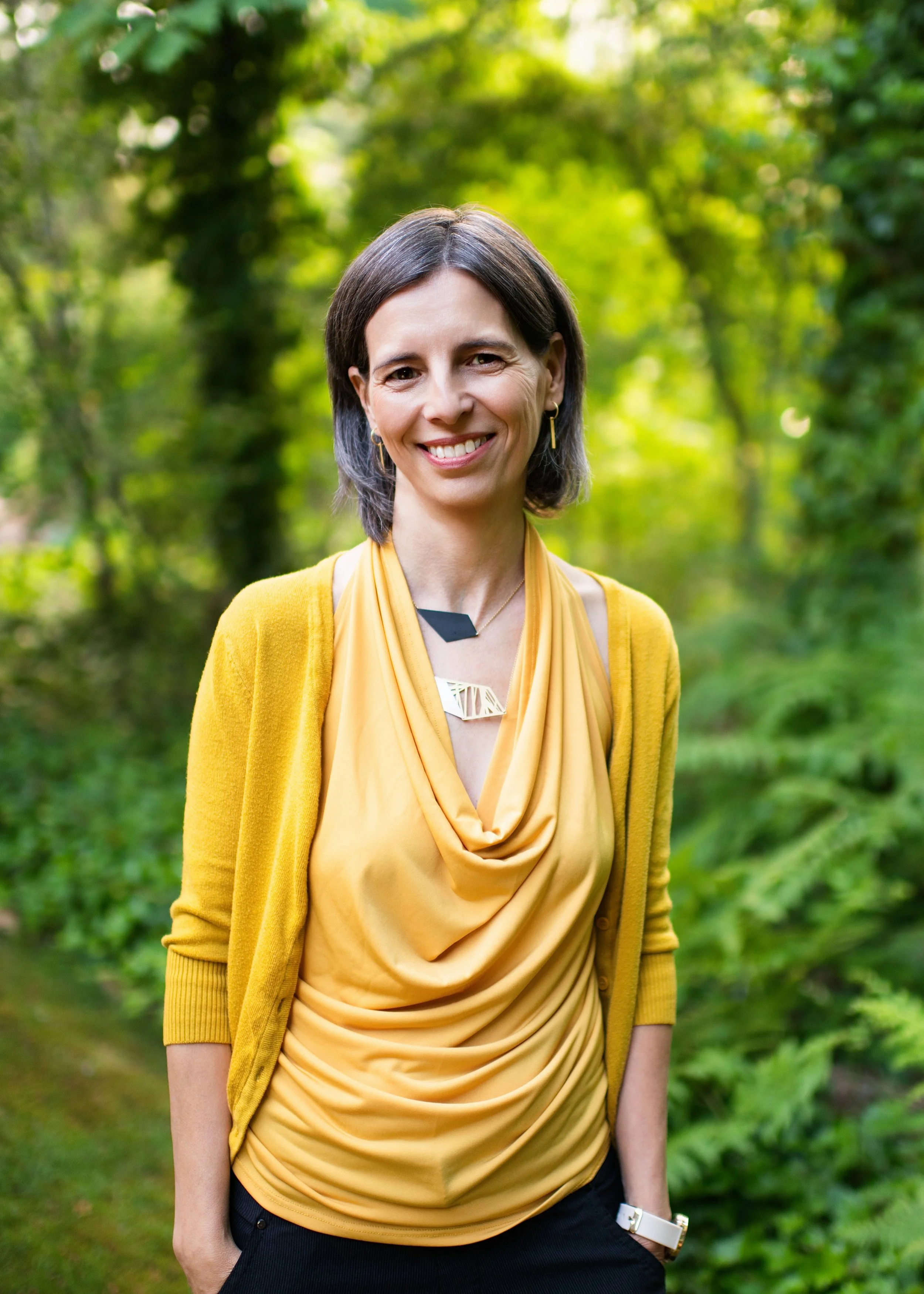 Close-up of a woman with light brown hair, smiling, wearing gold earrings, outdoors with green foliage and purple flowers in the background.