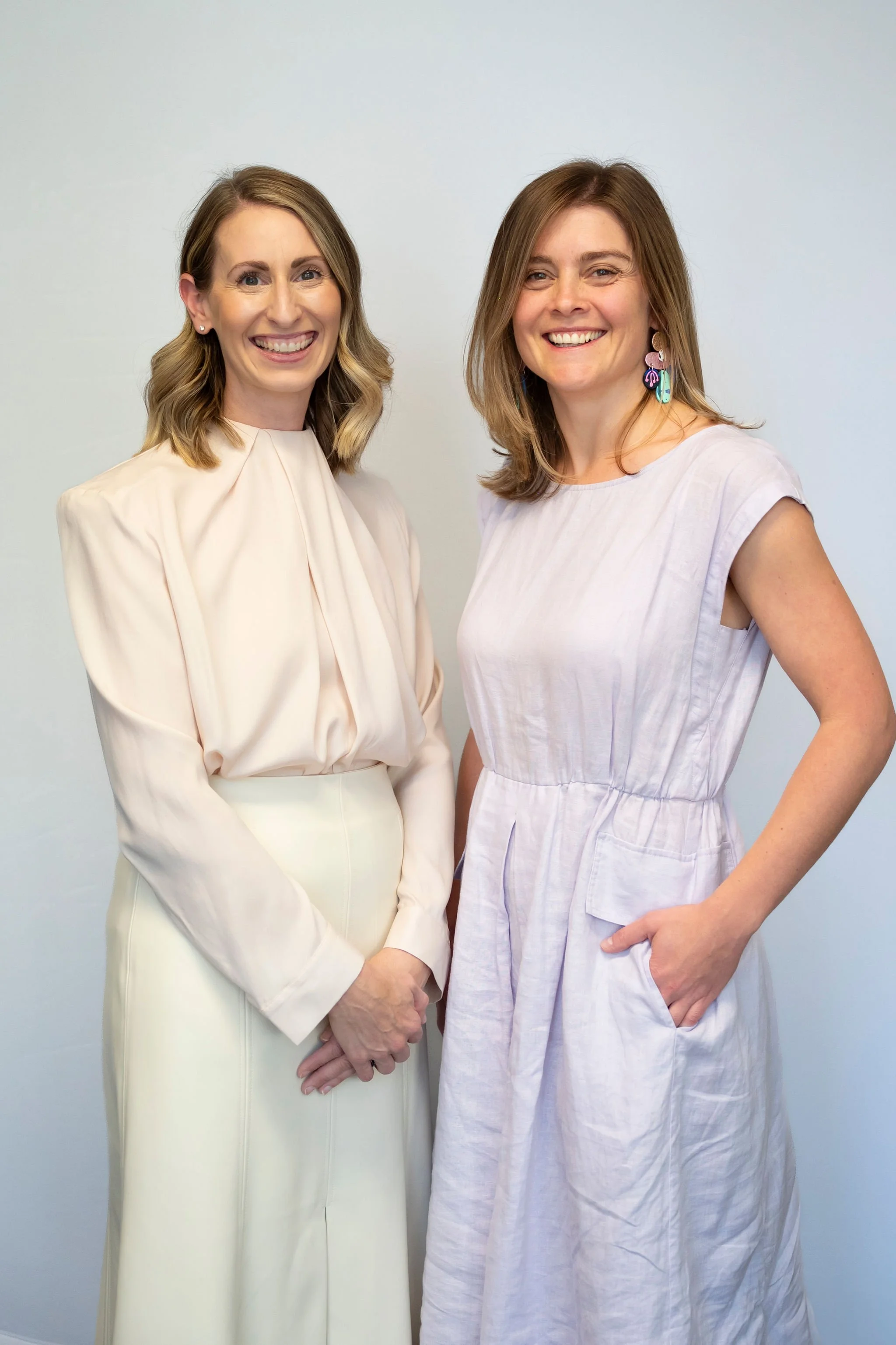 Two women standing side by side smiling, dressed in light-colored clothing, against a plain light background.
