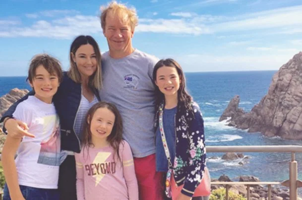 A family of five smiling near the ocean with rocky cliffs in the background.
