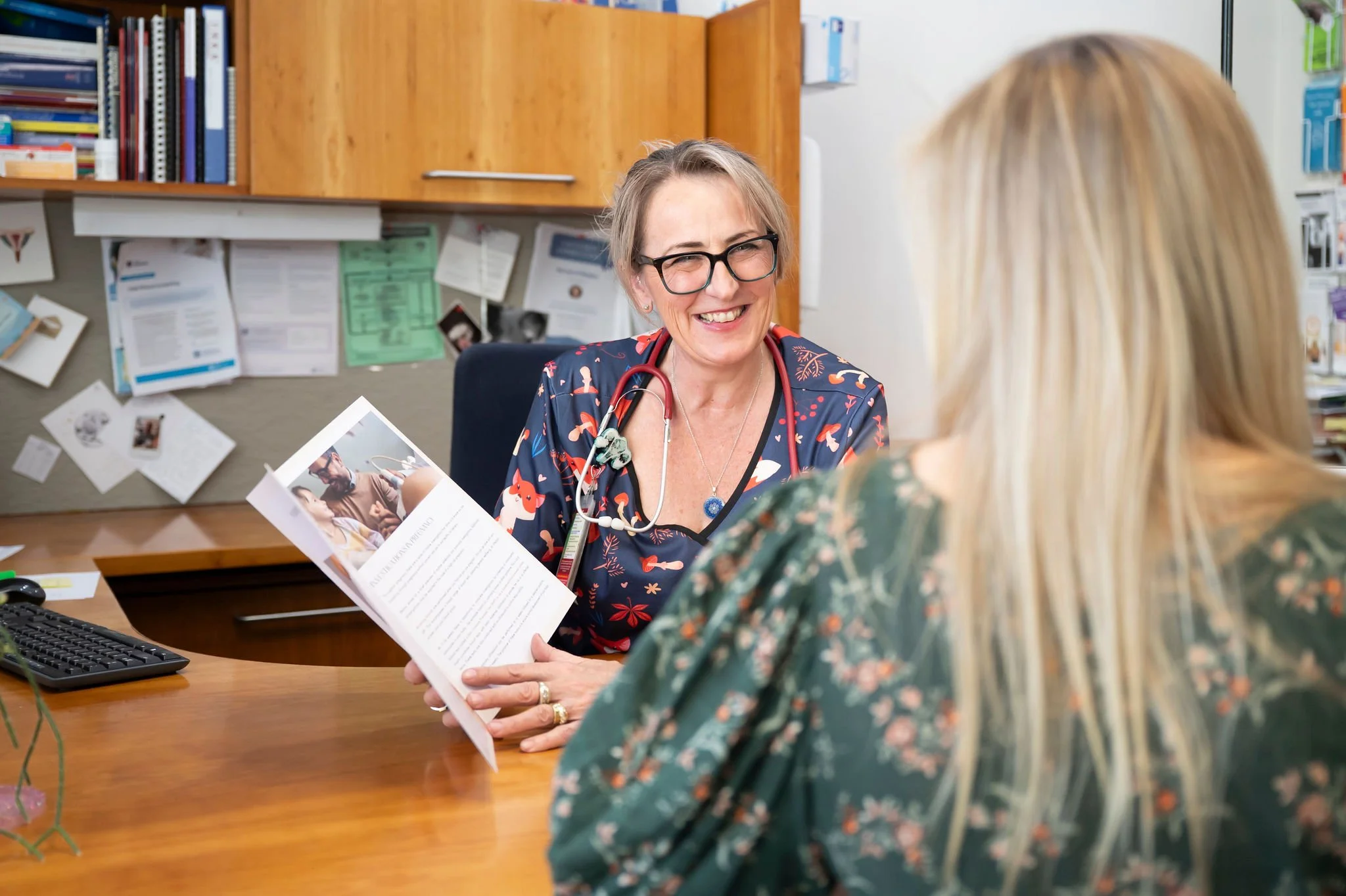 A female healthcare professional with glasses, blonde hair, and a stethoscope around her neck smiling and talking to a patient with blonde hair. They are in a medical office with bookshelves, papers, and a computer on the desk.