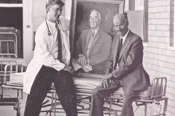 Two men, one in a doctor's coat and stethoscope, and the other in a suit, sitting and standing beside a framed portrait of a man, outdoors near a brick building.