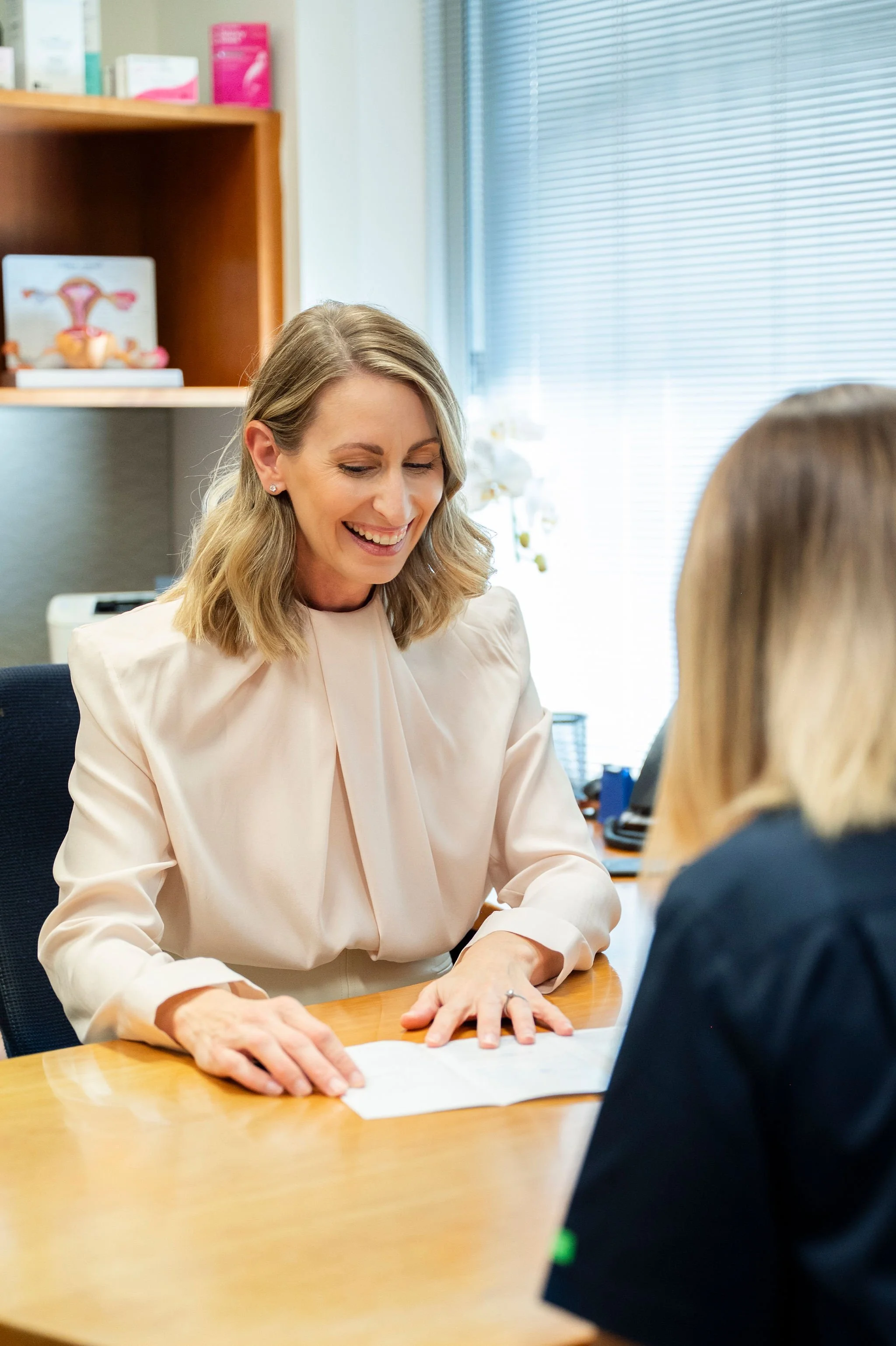 A woman with blonde hair and a light-colored blouse smiling, sitting at a desk, engaging in conversation with another person whose back is visible. The setting appears to be an office with shelves and closed blinds in the background.