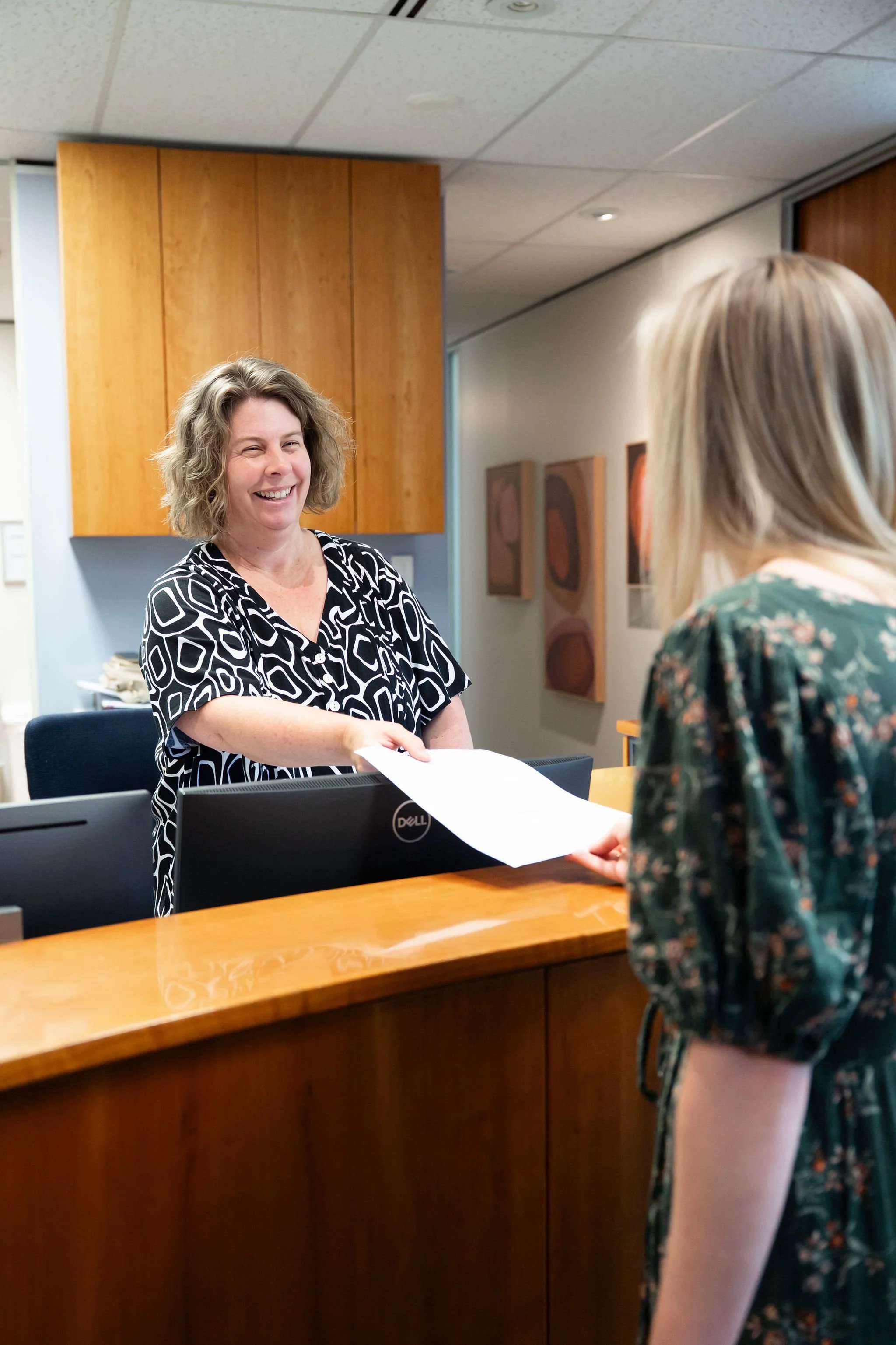 A woman with shoulder-length wavy hair in a patterned black and white shirt smiling and handing a piece of paper to another woman with blonde hair, wearing a floral top, at a reception desk in an office with wooden cabinets and abstract wall art.