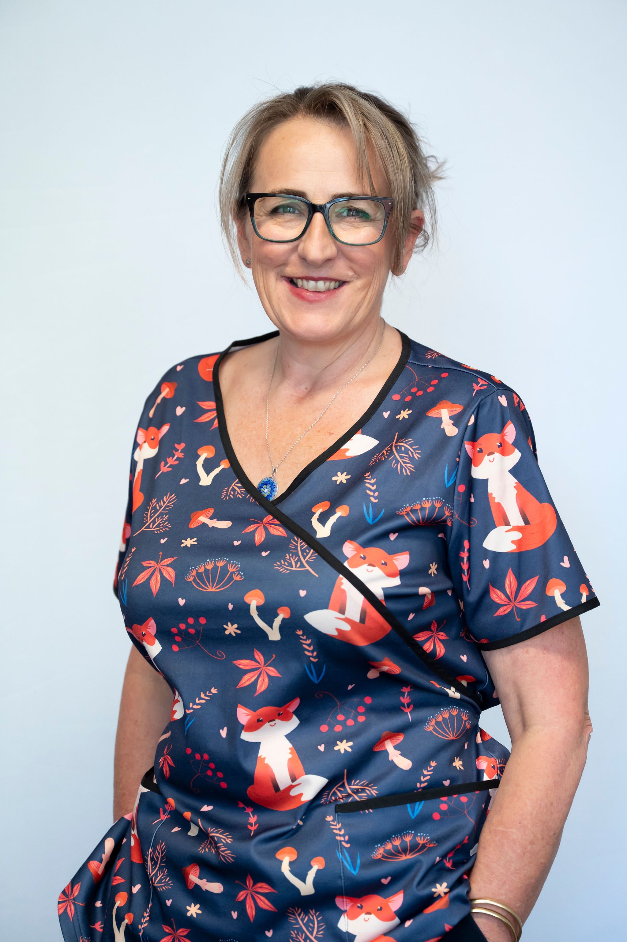 A smiling woman wearing glasses and a dark blue dress with a fox and autumn leaf pattern, standing against a plain white background.