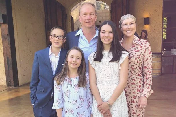 A family of five posing together indoors, with an archway and rustic decor in the background. The group includes a man, a woman, and three girls, all smiling.