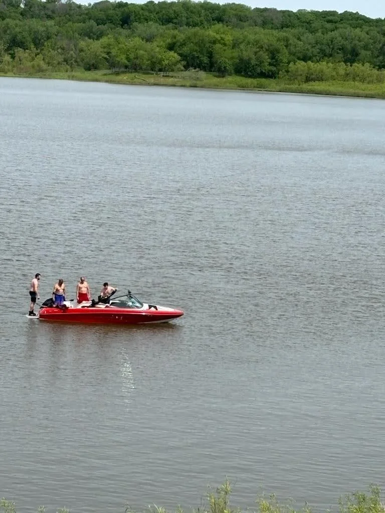 Four people on a red boat in a lake during daytime, with forested shoreline in the background.