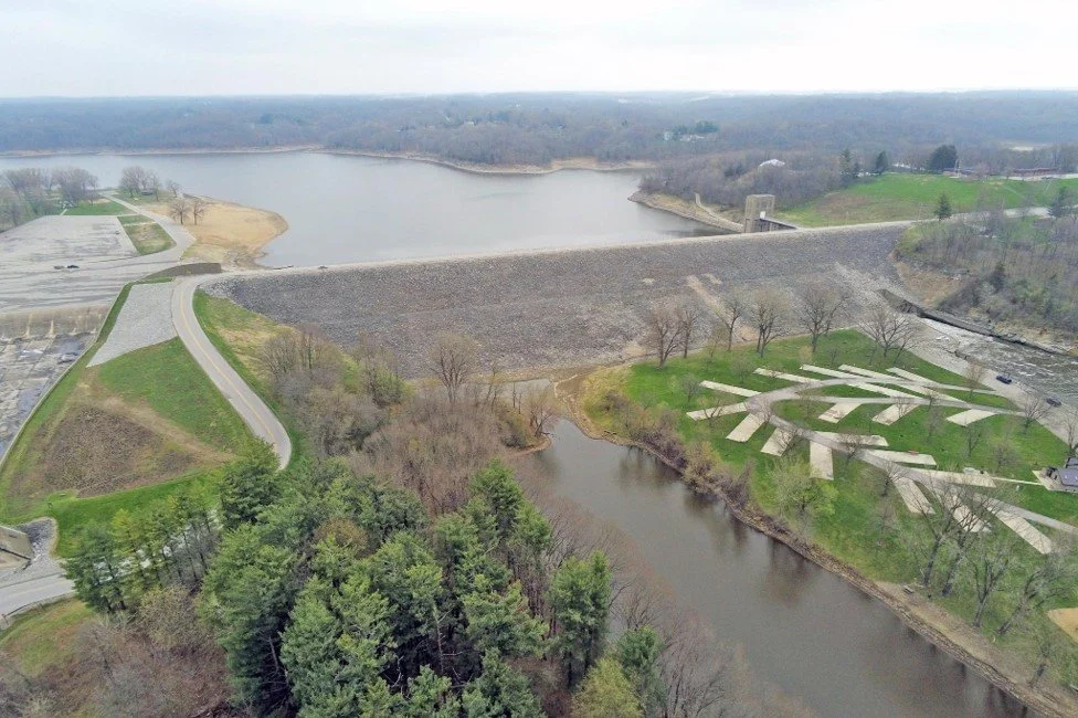 Aerial view of a dam with water reservoir, spillway with water flowing, surrounding trees, grassy areas, and concrete pathways with geometric design.