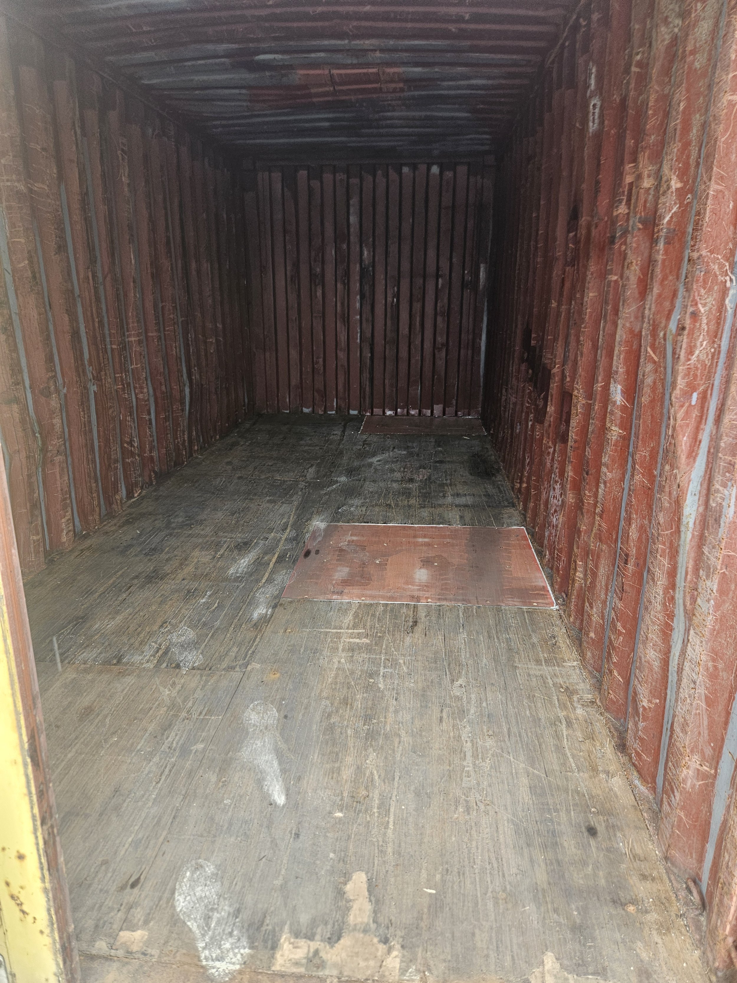 Empty and unfinished interior of a shipping container with wooden floor, and metal ceiling and walls, with you standing at the entrance.