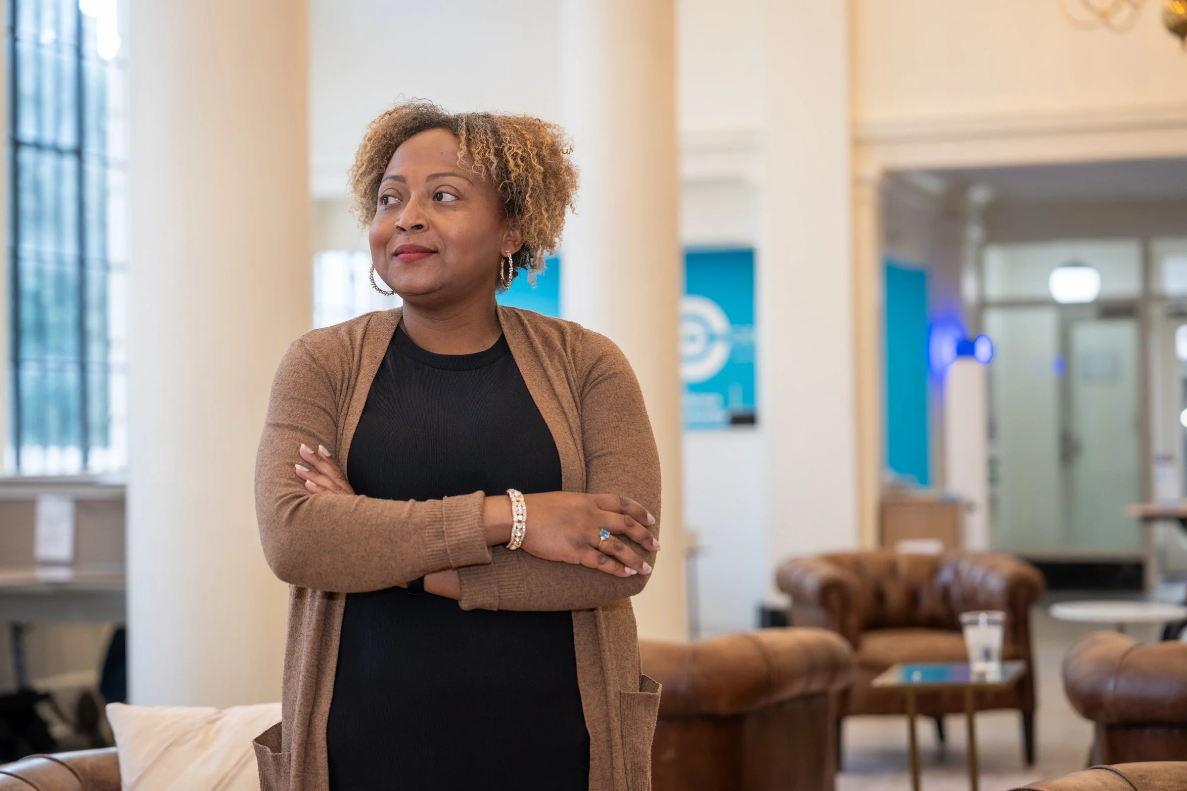 A woman with curly blonde hair wearing a black dress and brown cardigan standing with arms crossed in a lobby or waiting area with leather sofas, a glass of water, and windows.