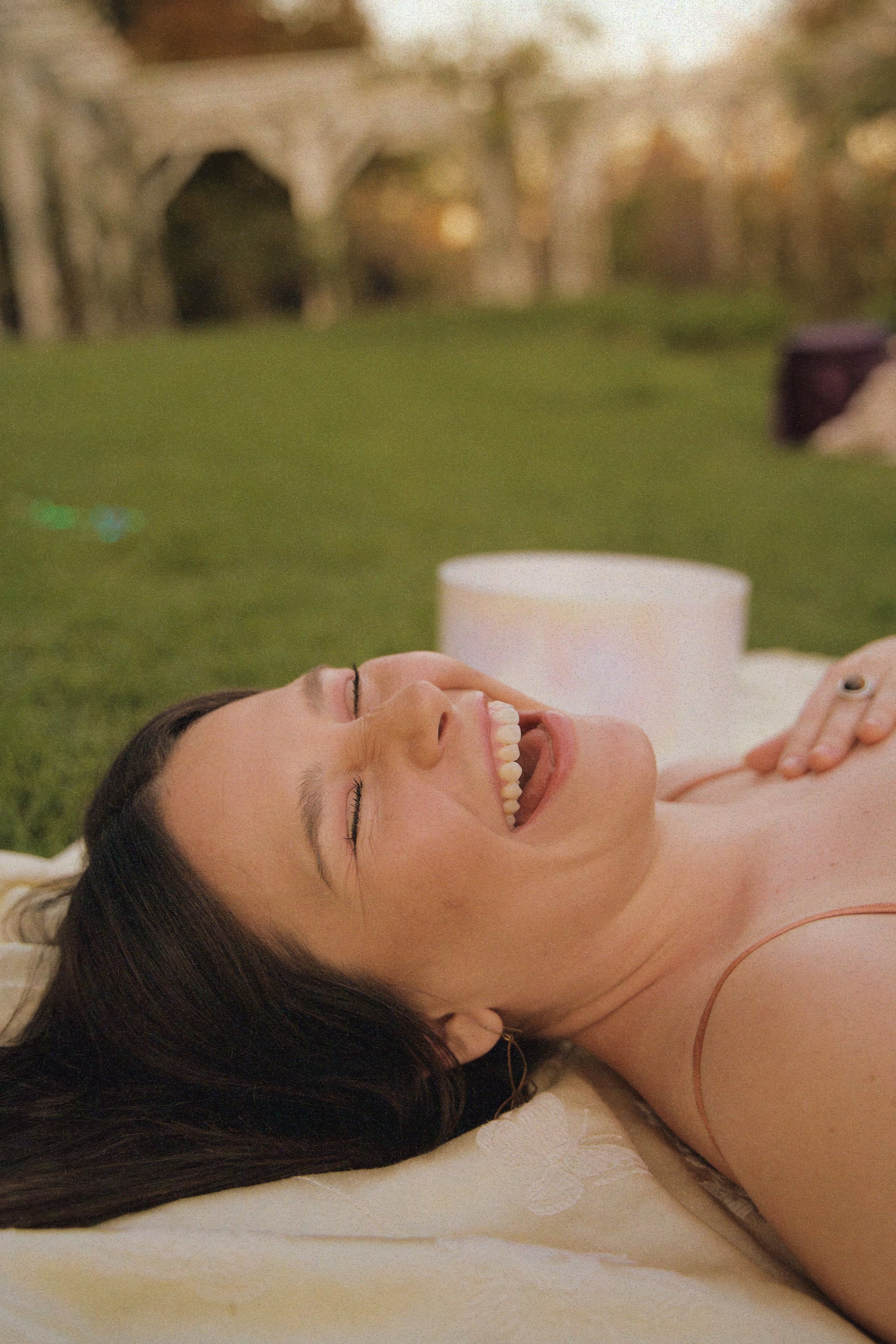 A woman with long dark hair lying on a blanket outdoors, laughing joyfully, with a music speaker nearby.