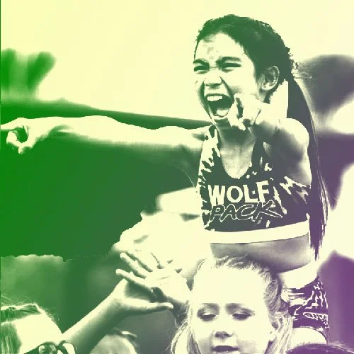 A young girl is pointing and shouting, with an enthusiastic expression, while another child is touching her arm and looking on. The background features natural scenery. Pack Athletics