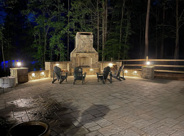 Nighttime outdoor patio with a stone fireplace, four black Adirondack chairs arranged around a firepit, and string lights illuminating the patio area, surrounded by trees.