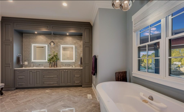 Modern bathroom with double vanity mirrors and a large corner bathtub by a window showing a sunrise or sunset.