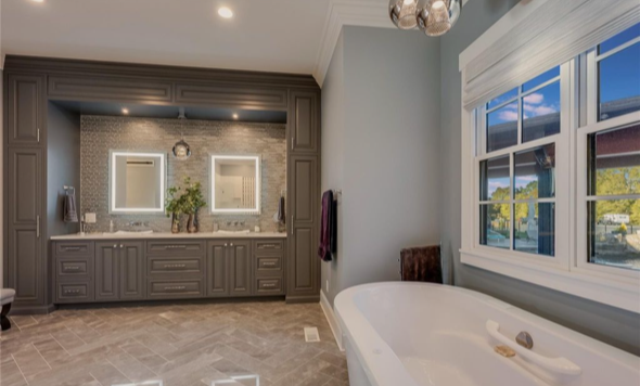 Bathroom with double vanity, illuminated mirrors, and a bathtub near a window showing greenery outside.