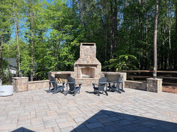 Outdoor patio area with stone fireplace, four black Adirondack chairs arranged around it, surrounded by a low stone wall, with a wooded background.