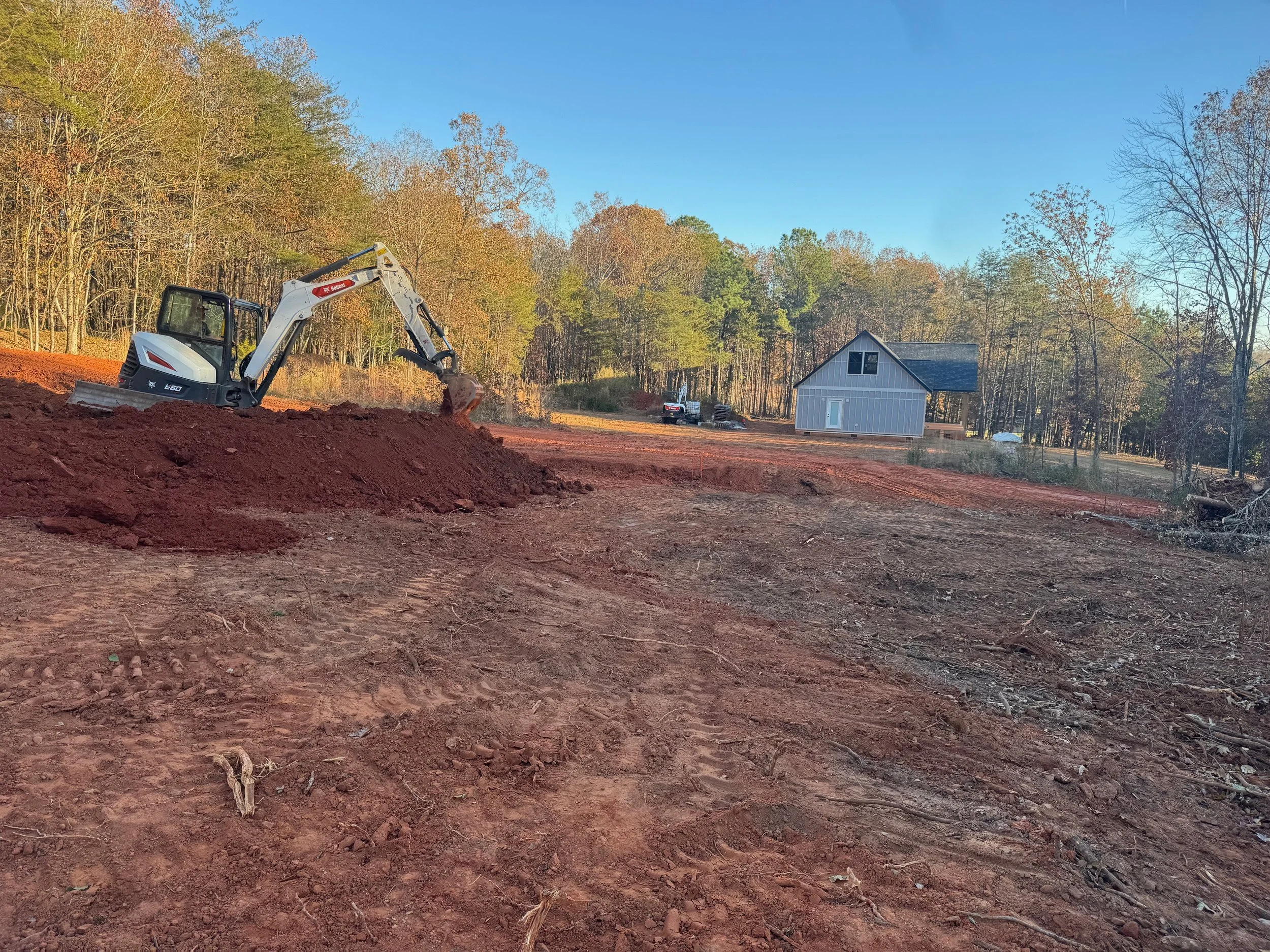 Construction site with a small excavator moving dirt, cleared land, and a house in the background amidst trees.
