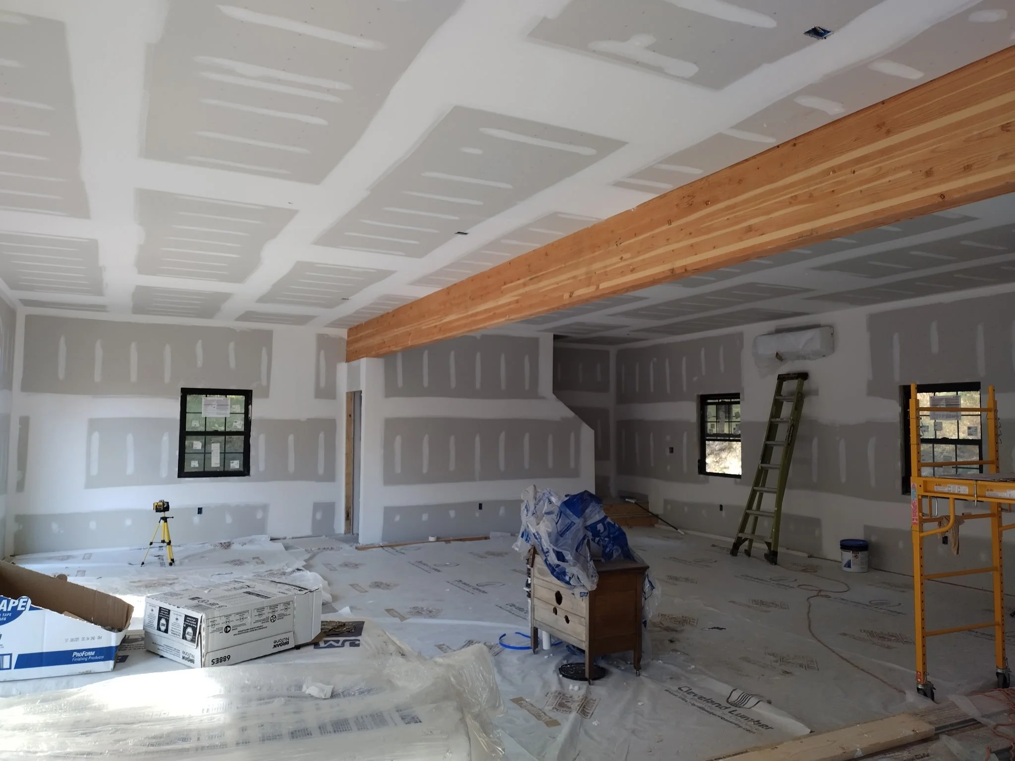 Interior of a room under construction with drywall walls and ceiling, a wooden beam across the ceiling, ladder, toolbox, and construction materials.