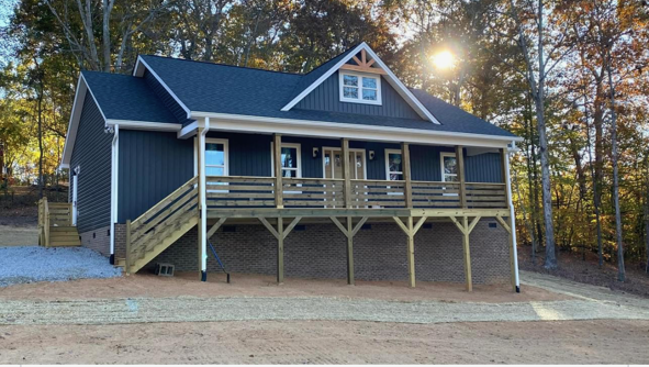 Newly built dark blue house with front porch, wooden railing, stairway, surrounded by trees, during sunset.
