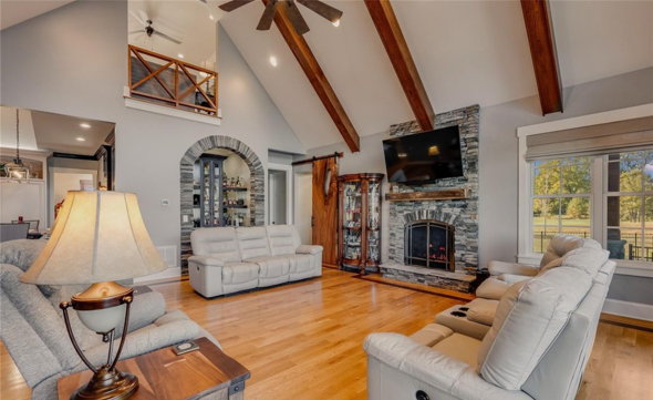 Living room with vaulted ceiling, exposed wooden beams, stone fireplace, flat-screen TV, and large windows with view of outdoors.