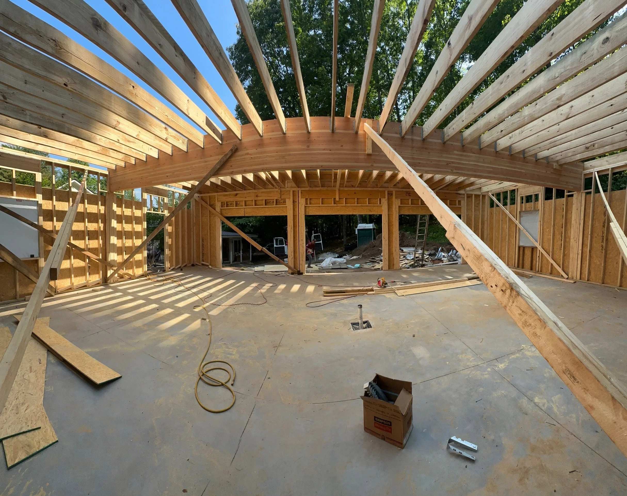 Interior of a house under construction with wooden framing and sunlight casting shadows through the beams.
