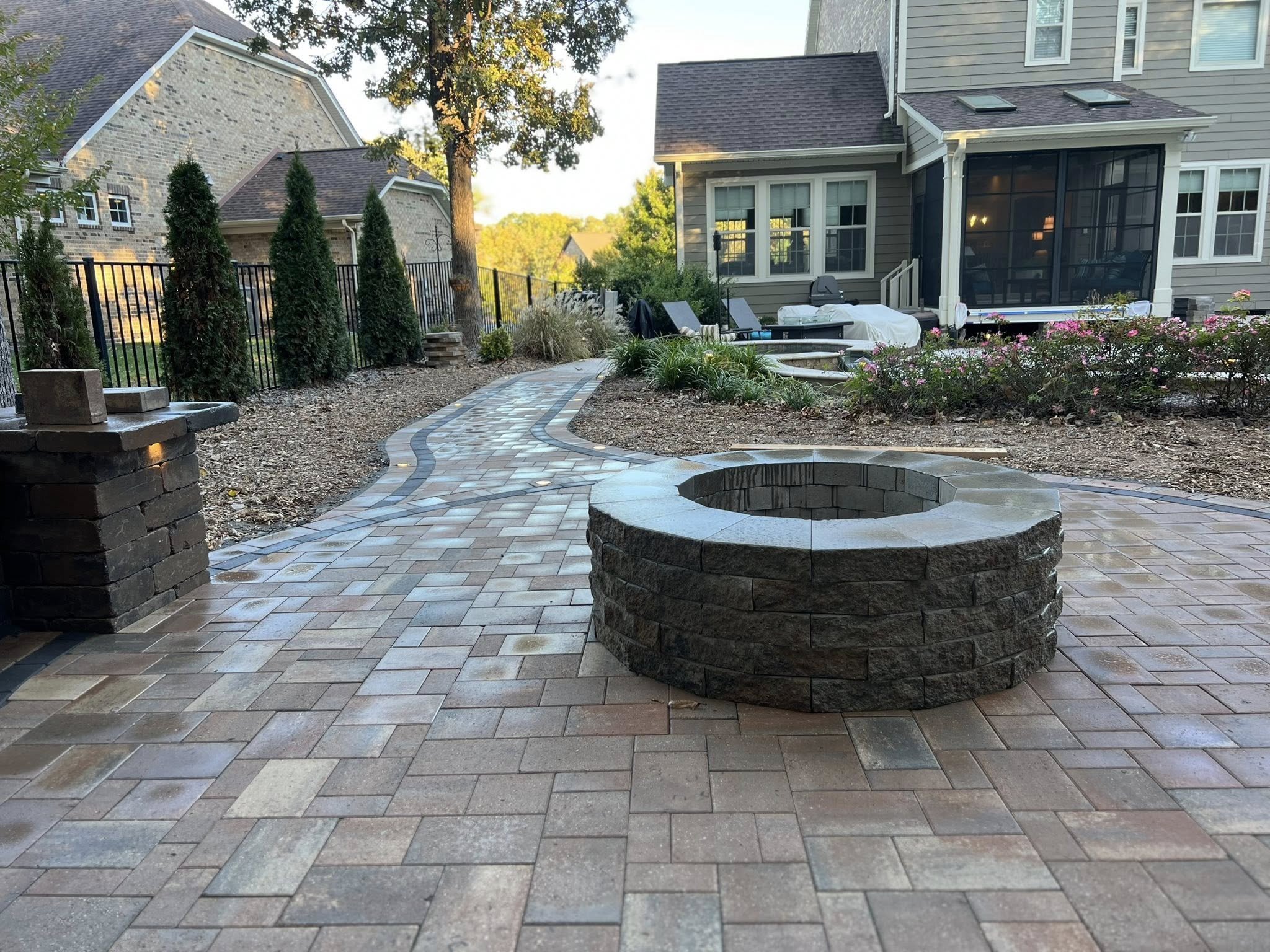 A backyard patio with a winding brick pathway, a circular stone fire pit, and a house with a screened porch.