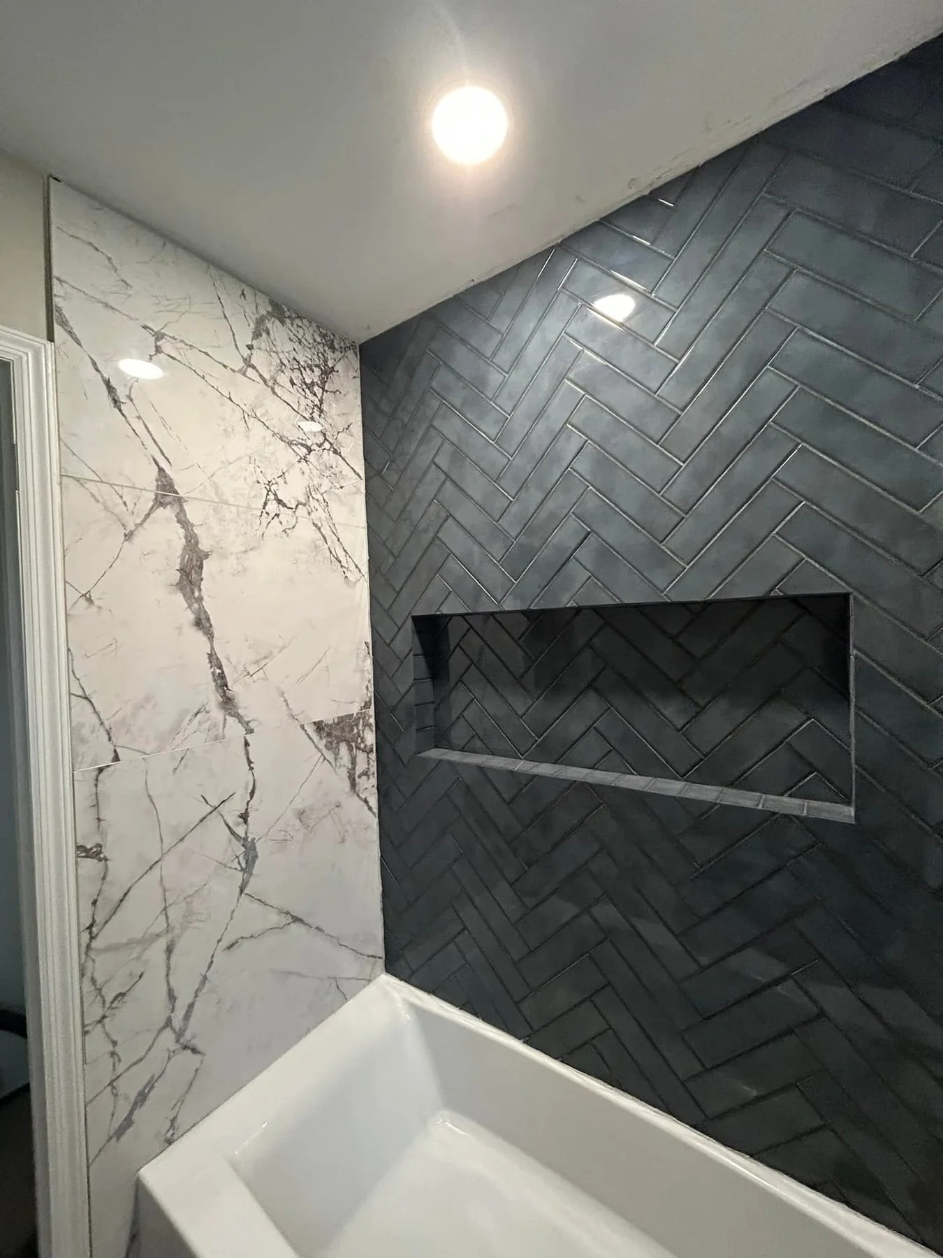 Close-up of a bathroom shower area with two different wall tiles: white marble with gray veining on the left and dark gray herringbone tile on the right, featuring a built-in niche for toiletries.