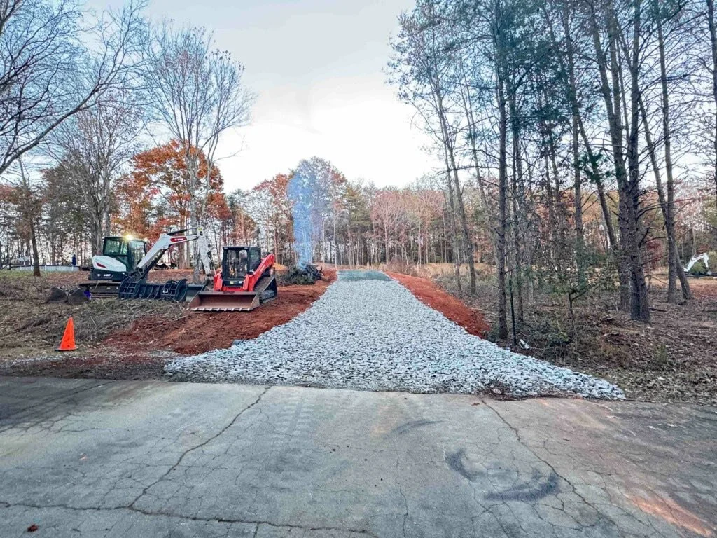 Construction site with gravel pathway being built through a wooded area, with construction equipment and orange safety cone visible.