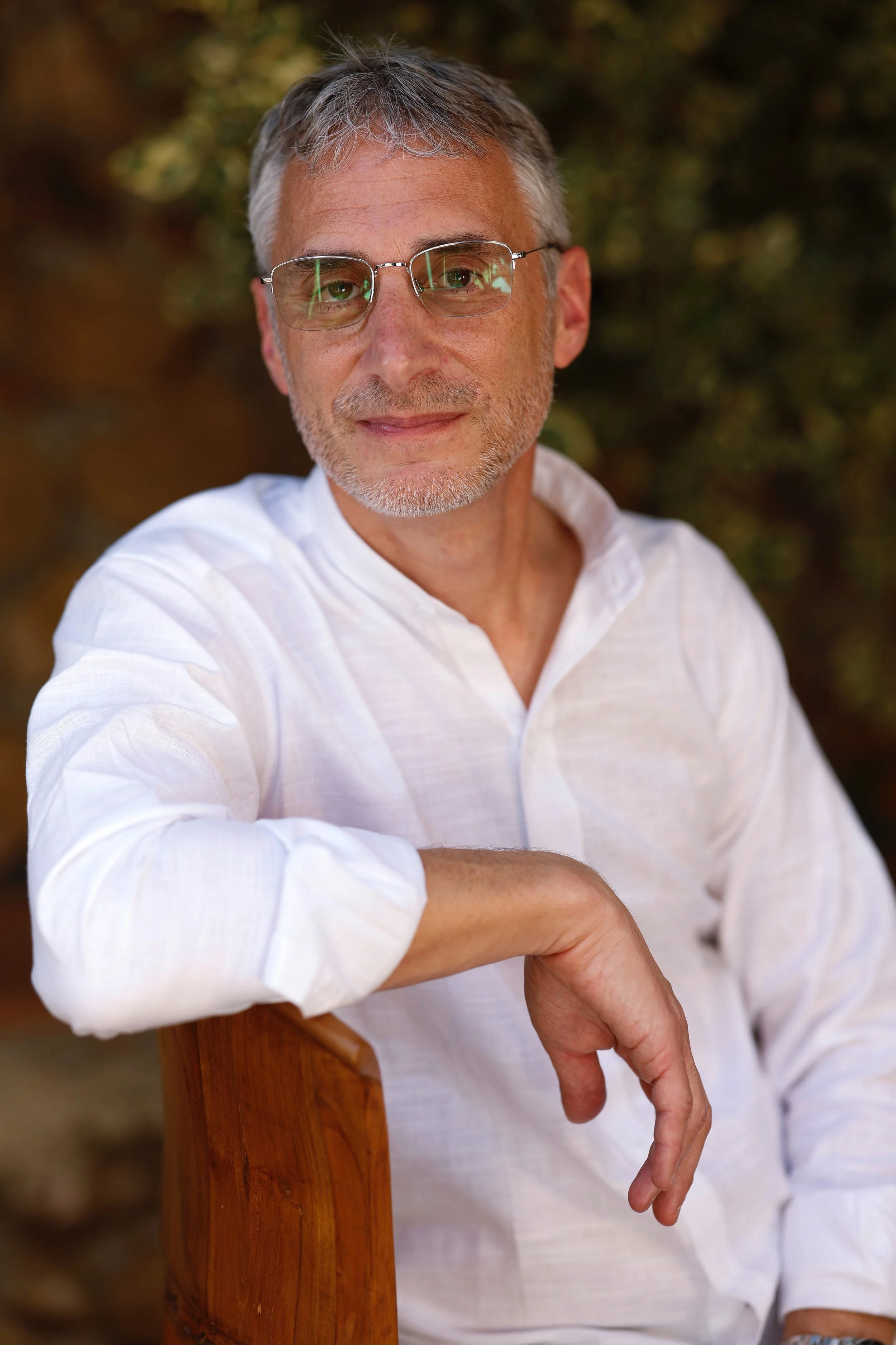 A middle-aged man with gray hair, glasses, and a beard, wearing a white shirt, sitting outdoors against a blurred background of greenery, looking at the camera with a slight smile.