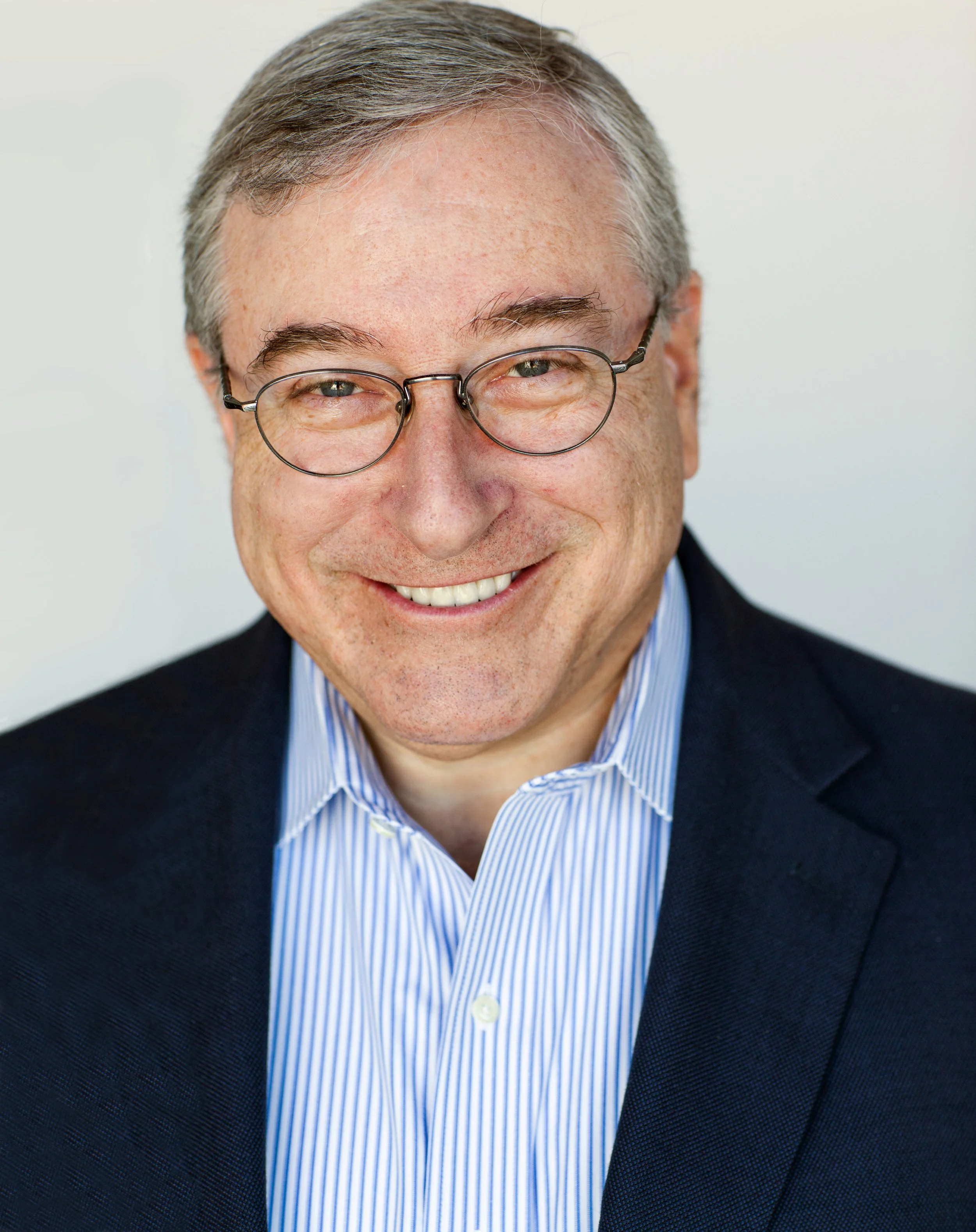 Headshot of a smiling middle-aged man with short gray hair, glasses, and wearing a navy blazer and light blue striped shirt.