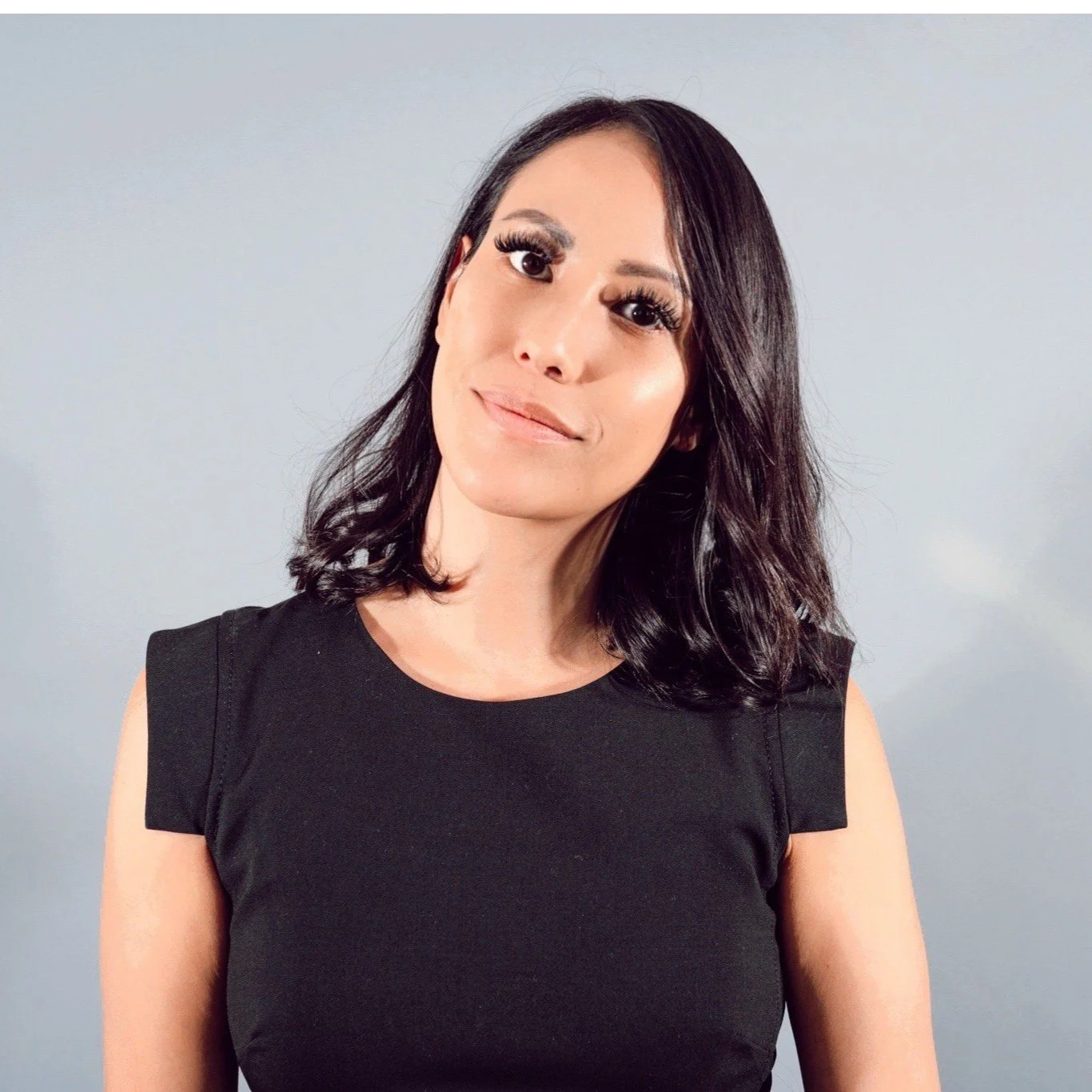 A woman with shoulder-length dark hair, wearing a black sleeveless top, standing against a light gray background, looking at the camera with a slight smile.