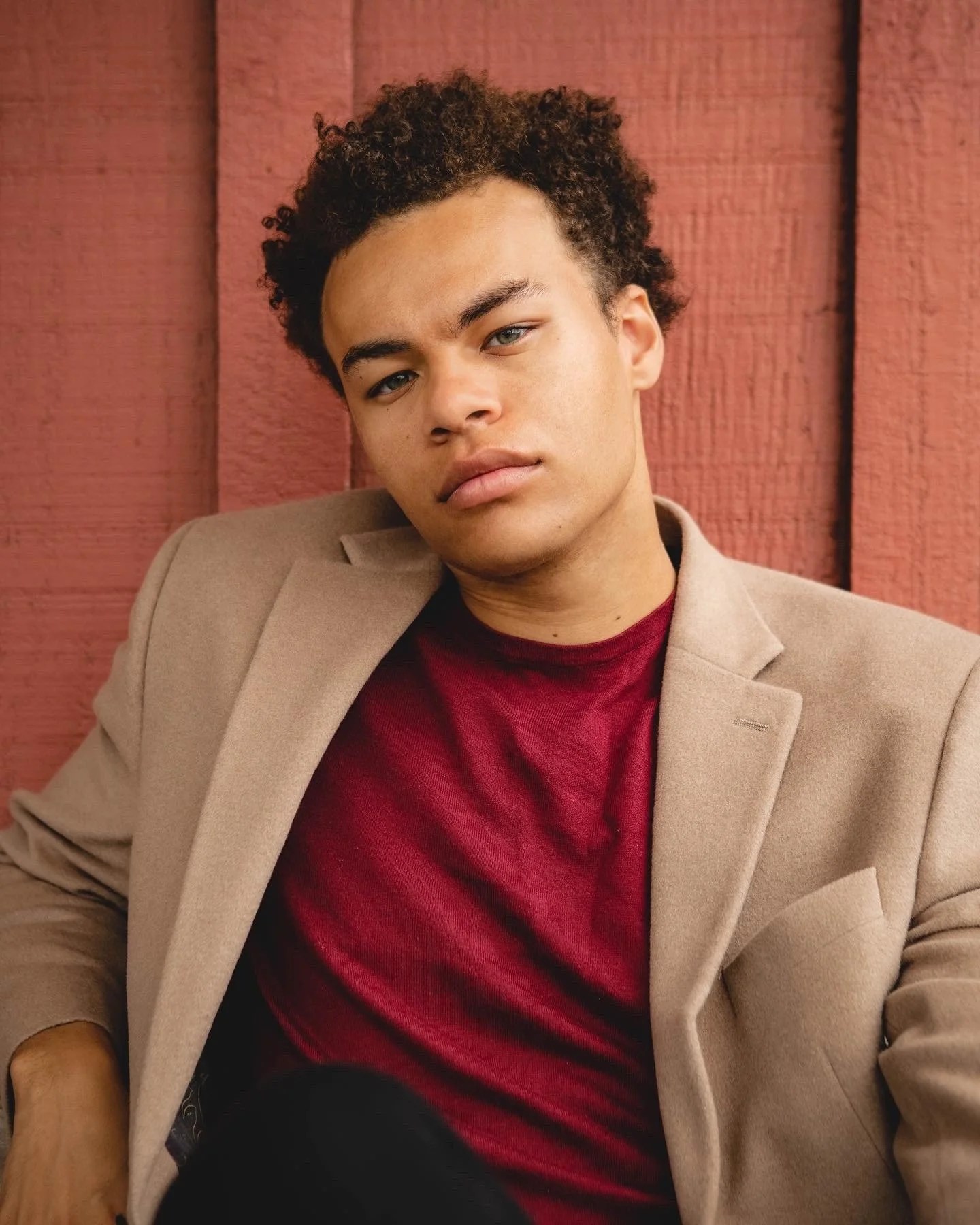 Young man with curly hair wearing a beige blazer and a red shirt, sitting against a red wooden wall.