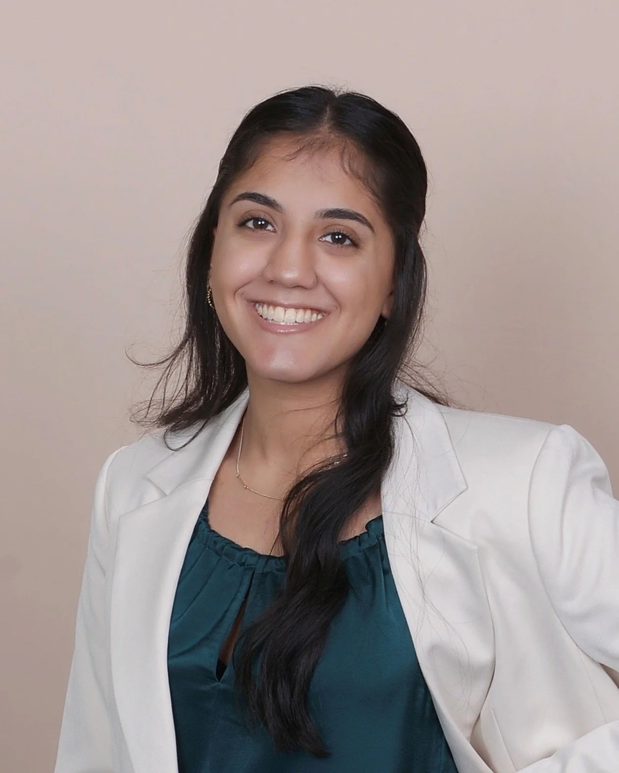A woman with long dark hair smiling, wearing a white blazer and a teal blouse, standing against a beige background.