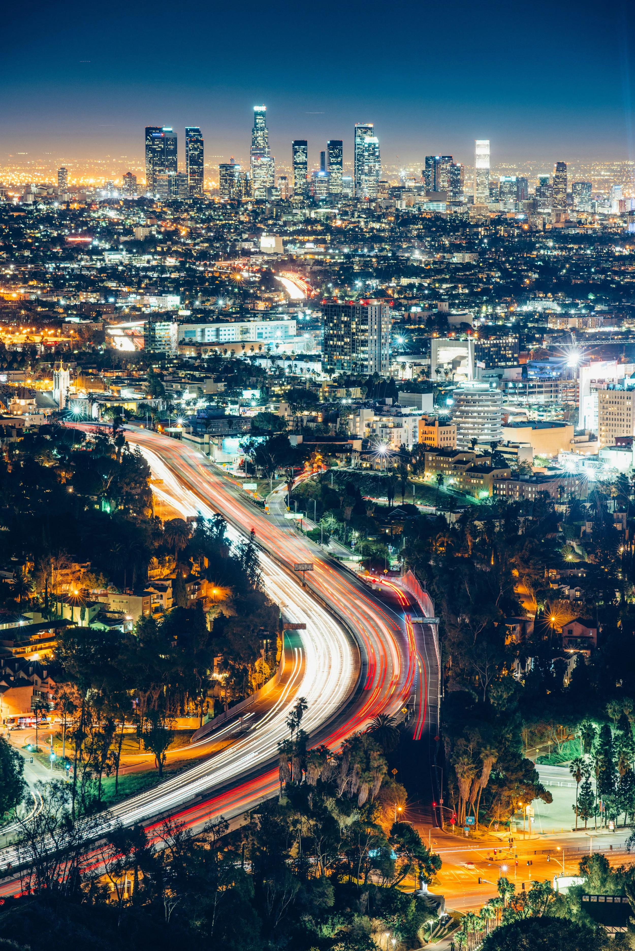 Nighttime view of a city skyline with tall illuminated skyscrapers and a busy highway with light trails from moving vehicles.