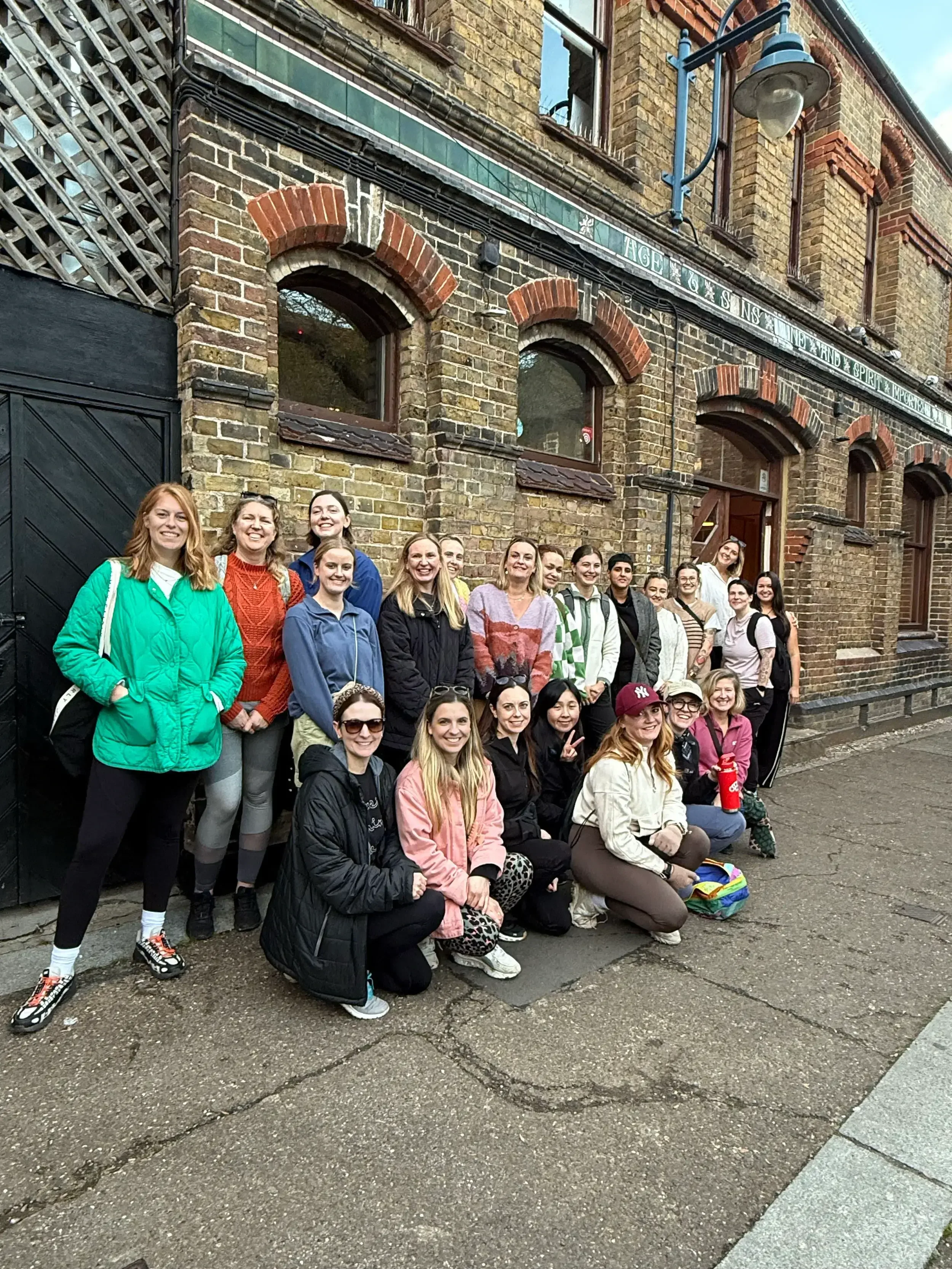 A large, diverse group of about twenty women posing for a group photo in front of a historic two-story brick building with arched windows. The women are dressed in casual hiking and autumn layers, including puffer jackets and fleeces.