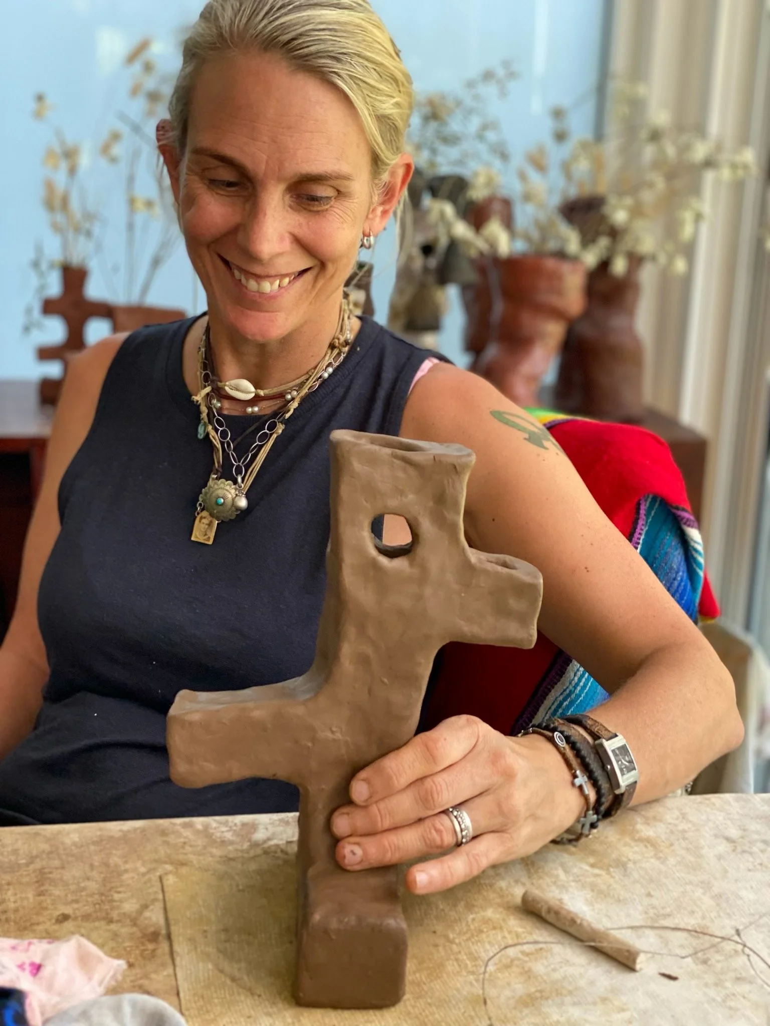 A woman with short blonde hair, wearing tattoos and layered necklaces, is smiling while holding a tan-colored ceramic sculpture of an abstract animal figure with a hole for an eye. She is sitting at a table with art supplies, in a room with vases of dried flowers and large windows.